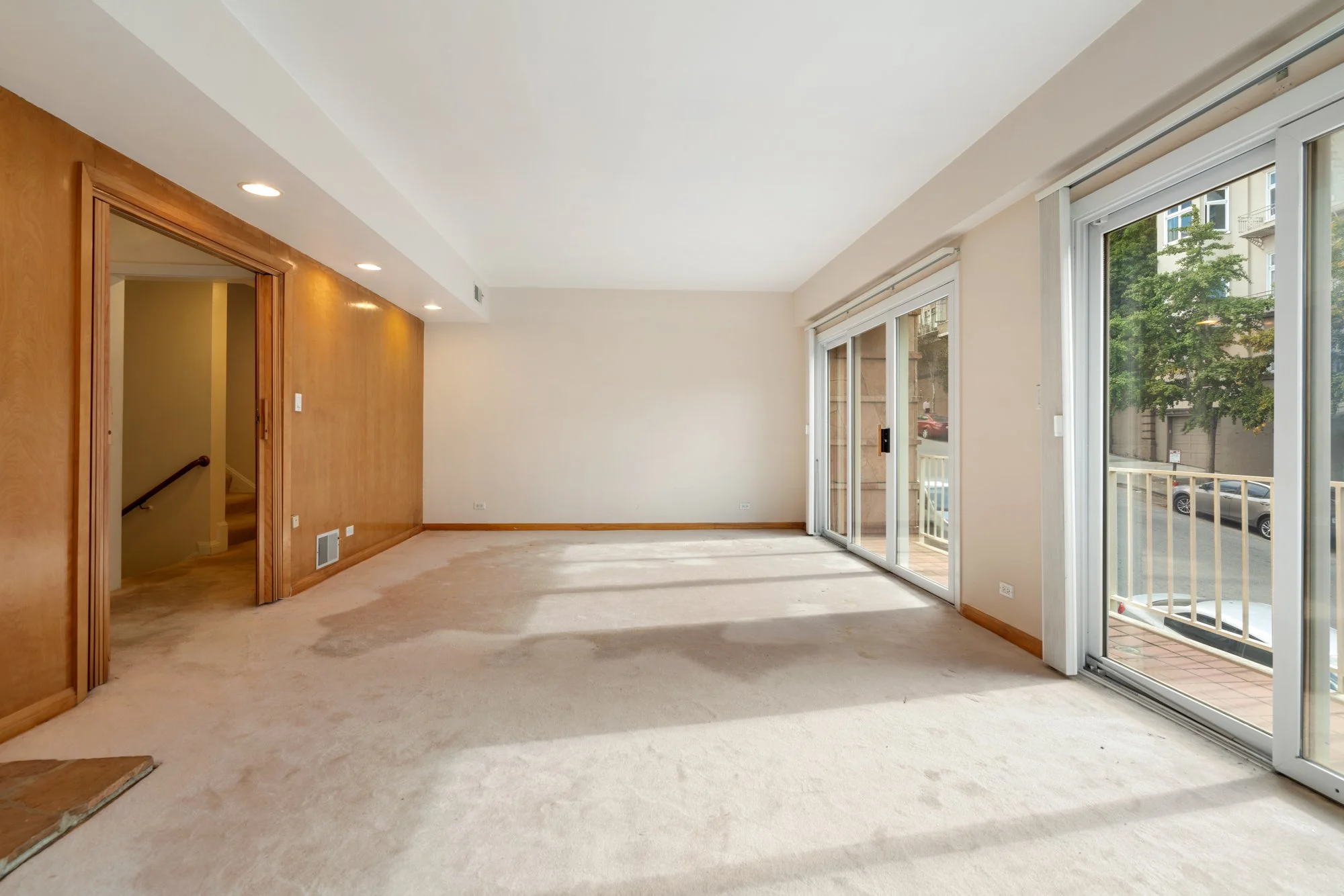 Empty living room with large sliding glass door leading to balcony, wood-paneled accent wall, beige carpet, and recessed ceiling lights.