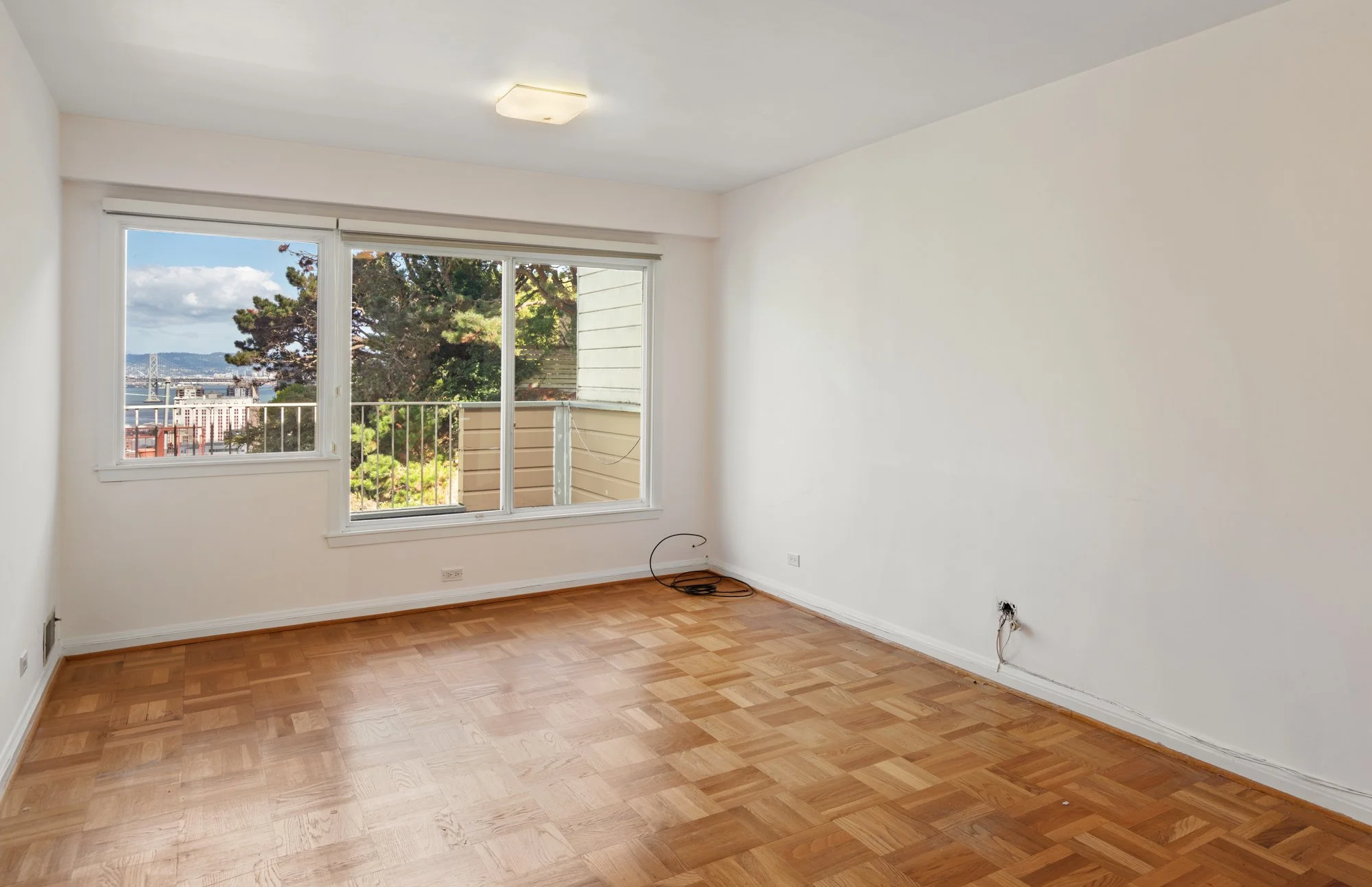 Empty room with hardwood floors, white walls, a ceiling light fixture, and large sliding glass doors leading to a balcony with a view of trees, water, and distant cityscape.