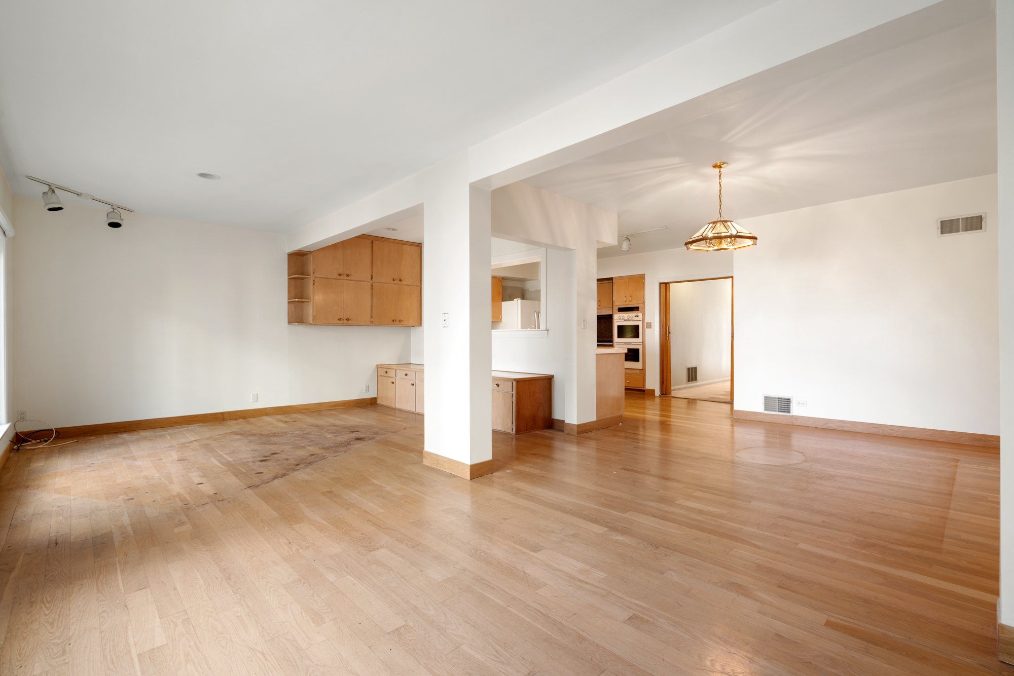 Empty living space with hardwood flooring, white walls, a ceiling with track lighting, and a hanging light fixture. Kitchen visible in the background with wooden cabinets and appliances.