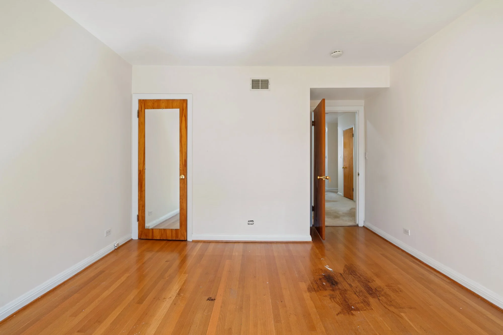 A room with hardwood floors, off-white walls, and two wooden doors partially open. One door has a full-length mirror. The floor shows some dark stains or discoloration.