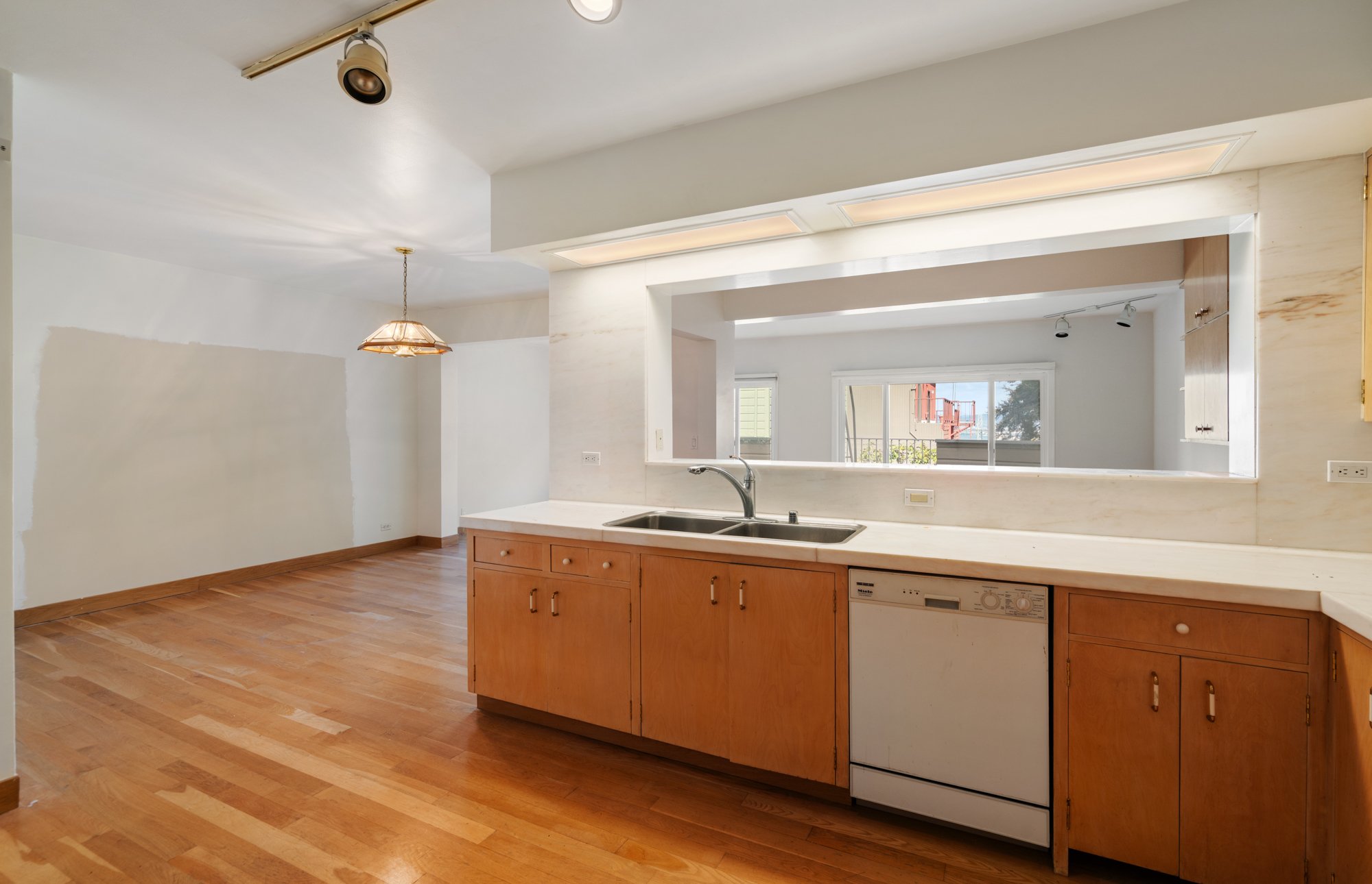 Living room and kitchen area with wooden flooring, white walls, a kitchen island with a sink, and large windows with a view of neighboring buildings and trees.