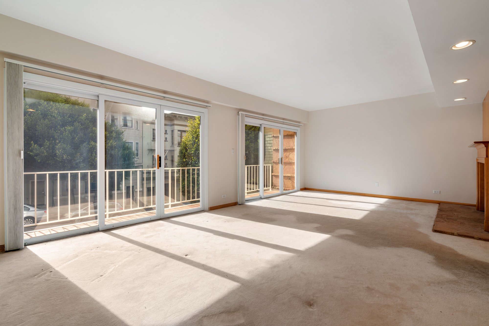 Empty room with large sliding glass doors leading to a balcony, sunlight casting shadows on the beige carpet, and trees visible outside.