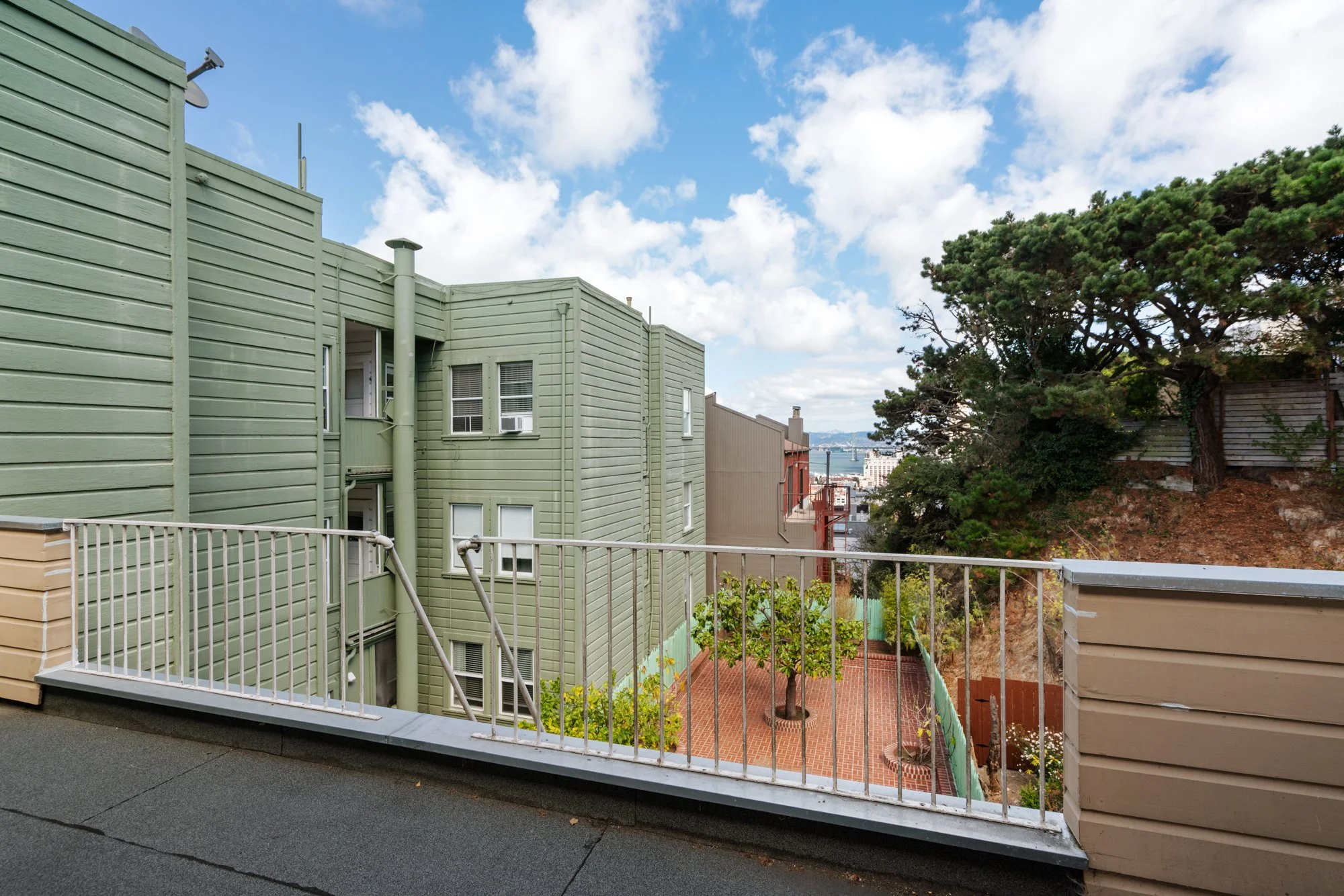 Apartment building with green siding viewed from a balcony patio, a metal railing, and a view of a cityscape and bay in the distance under a partly cloudy sky.