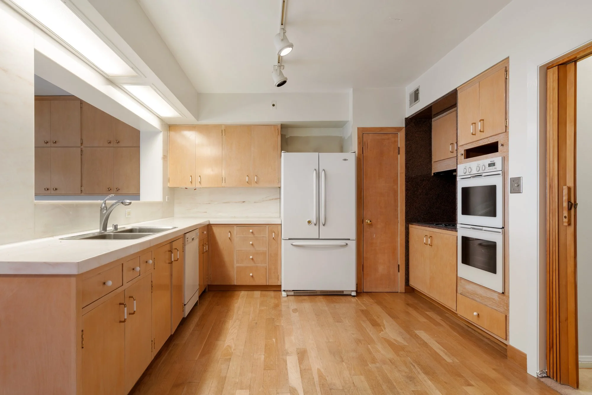 A kitchen with wooden cabinets, a white refrigerator, oven, and a countertop with a sink, wood flooring, and a door on the right side.