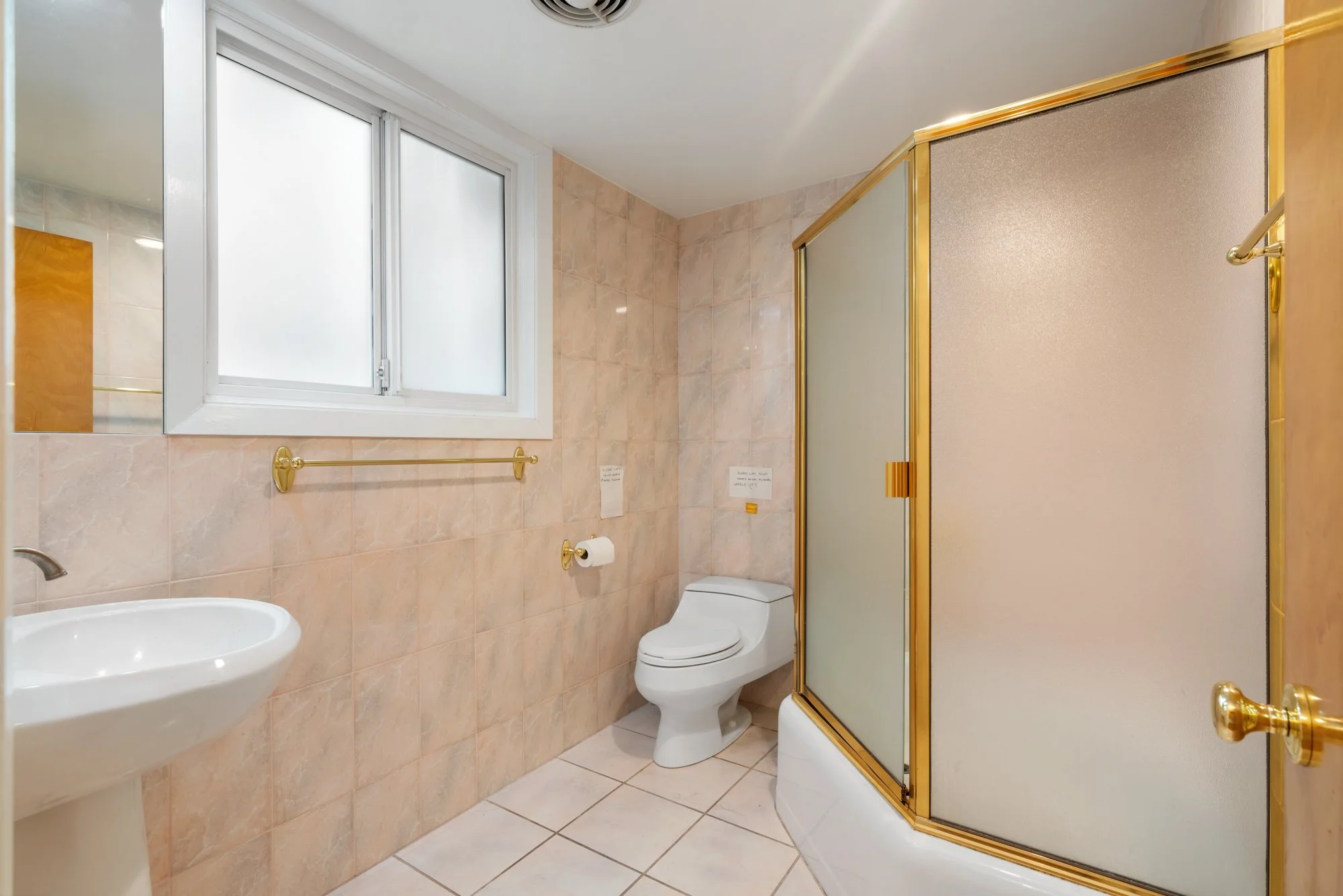 Bathroom with beige tile walls and floor, a white window, a toilet, a small sink, and a gold-framed shower with frosted glass door.