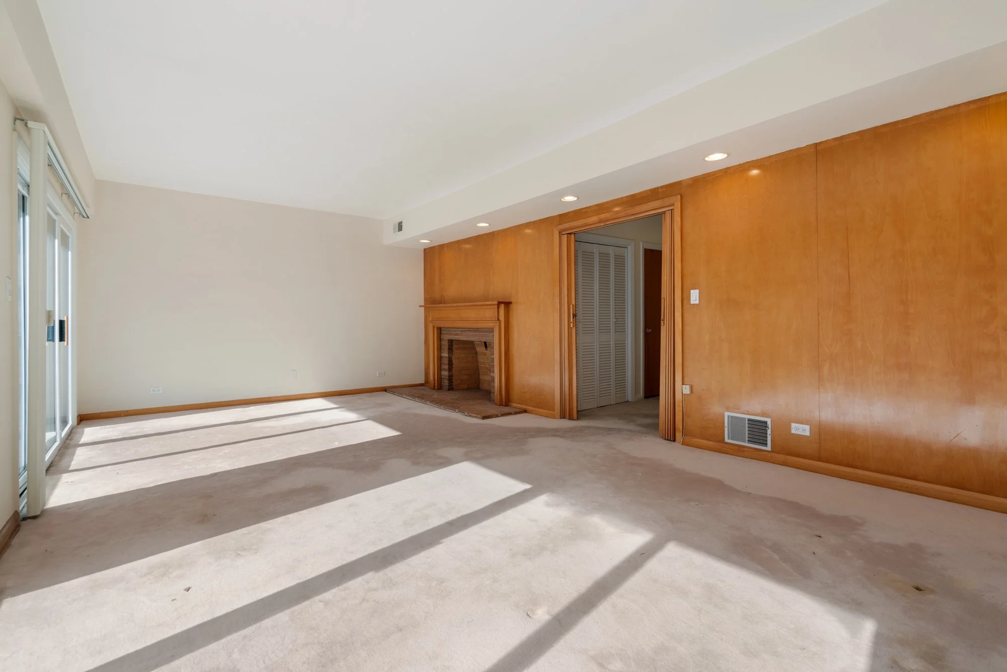 Empty living room with beige carpet, large sliding glass door casting sunlight and shadows, wood fireplace, and wood trim on walls.