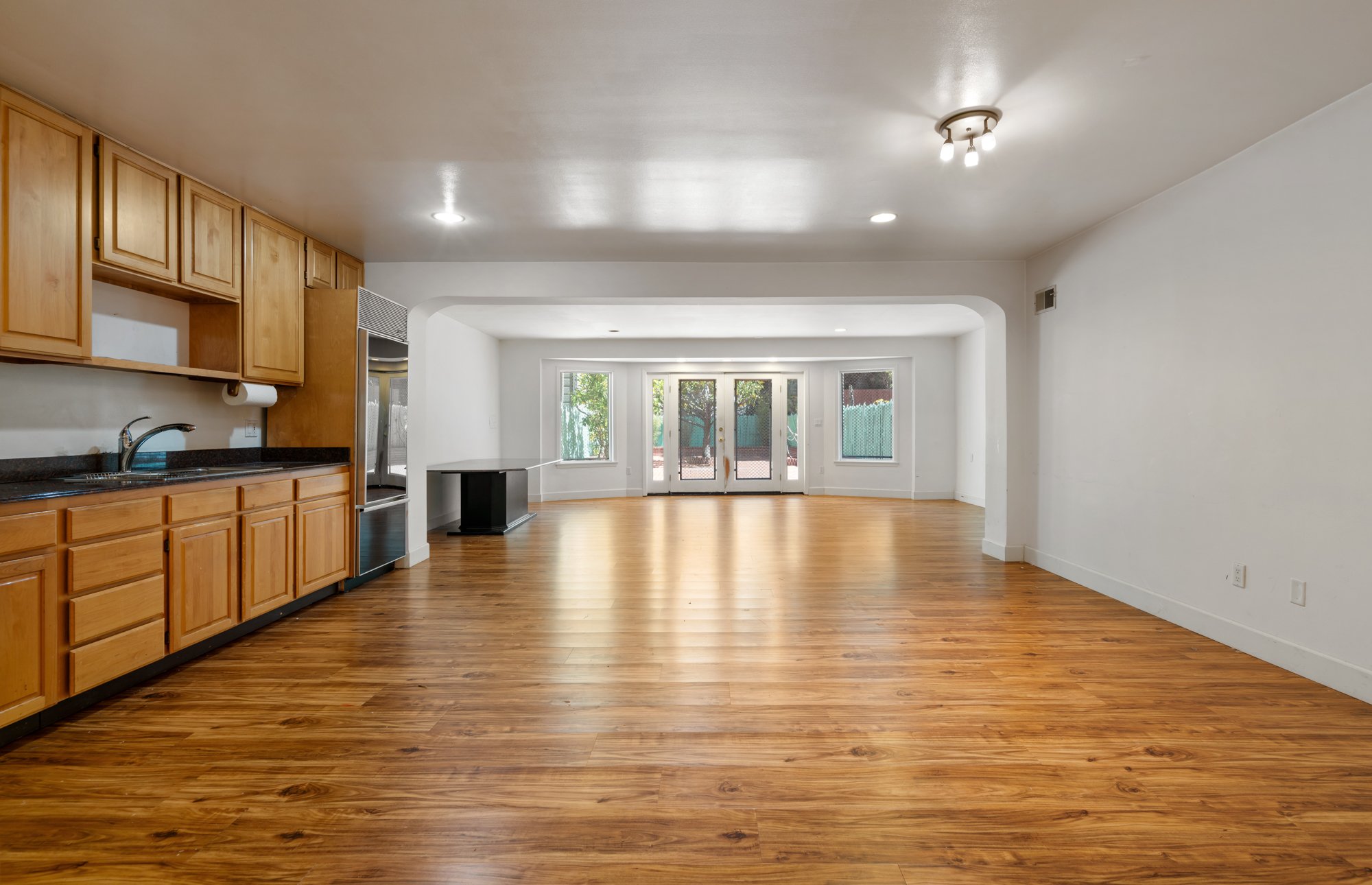 Open-concept kitchen and living area with wooden floors, light-colored wooden cabinets, large windows, and a sliding glass door leading outside.