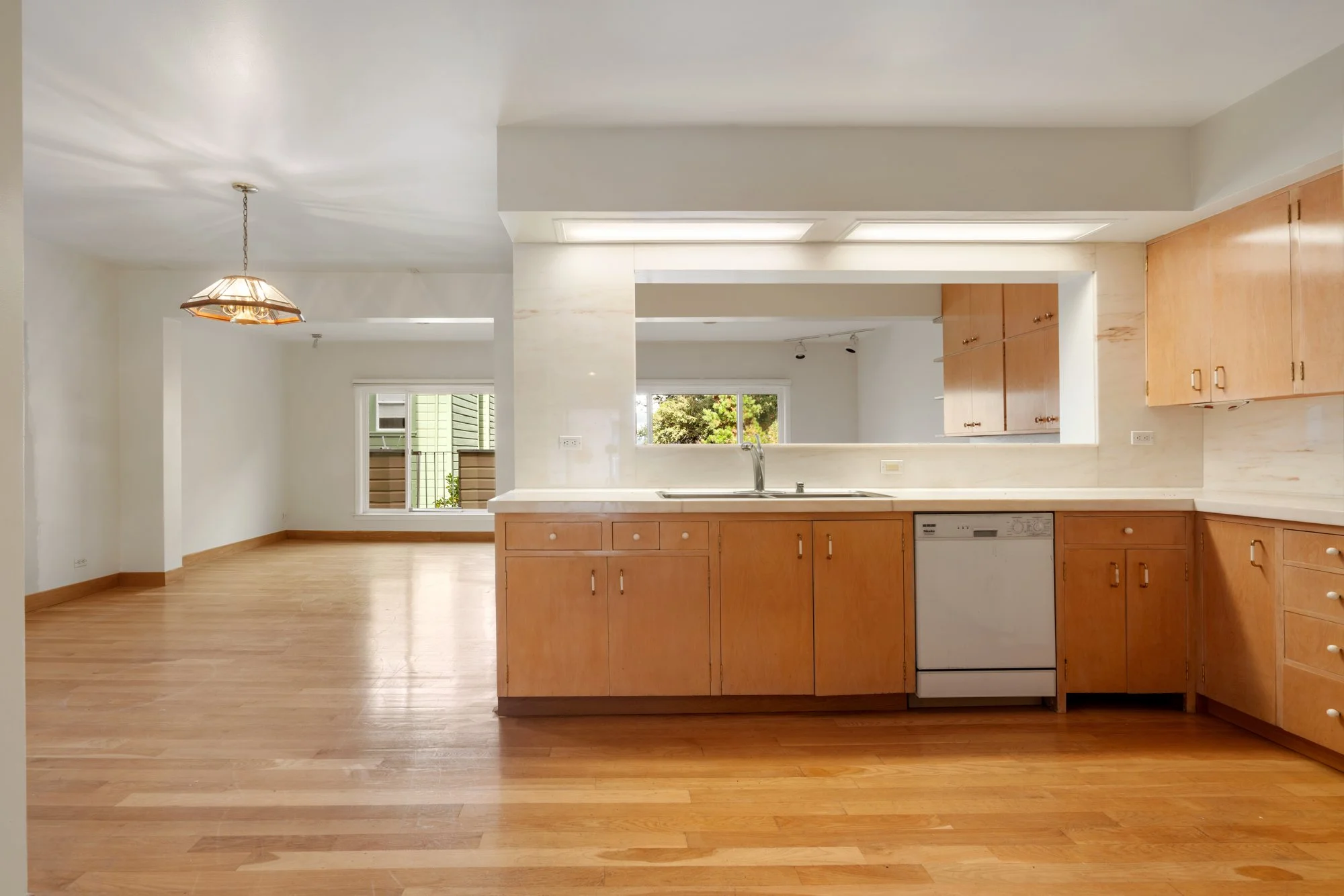 Open-concept kitchen with wooden cabinets, white countertops, and hardwood flooring, featuring a view into a living area with large windows and a pendant light