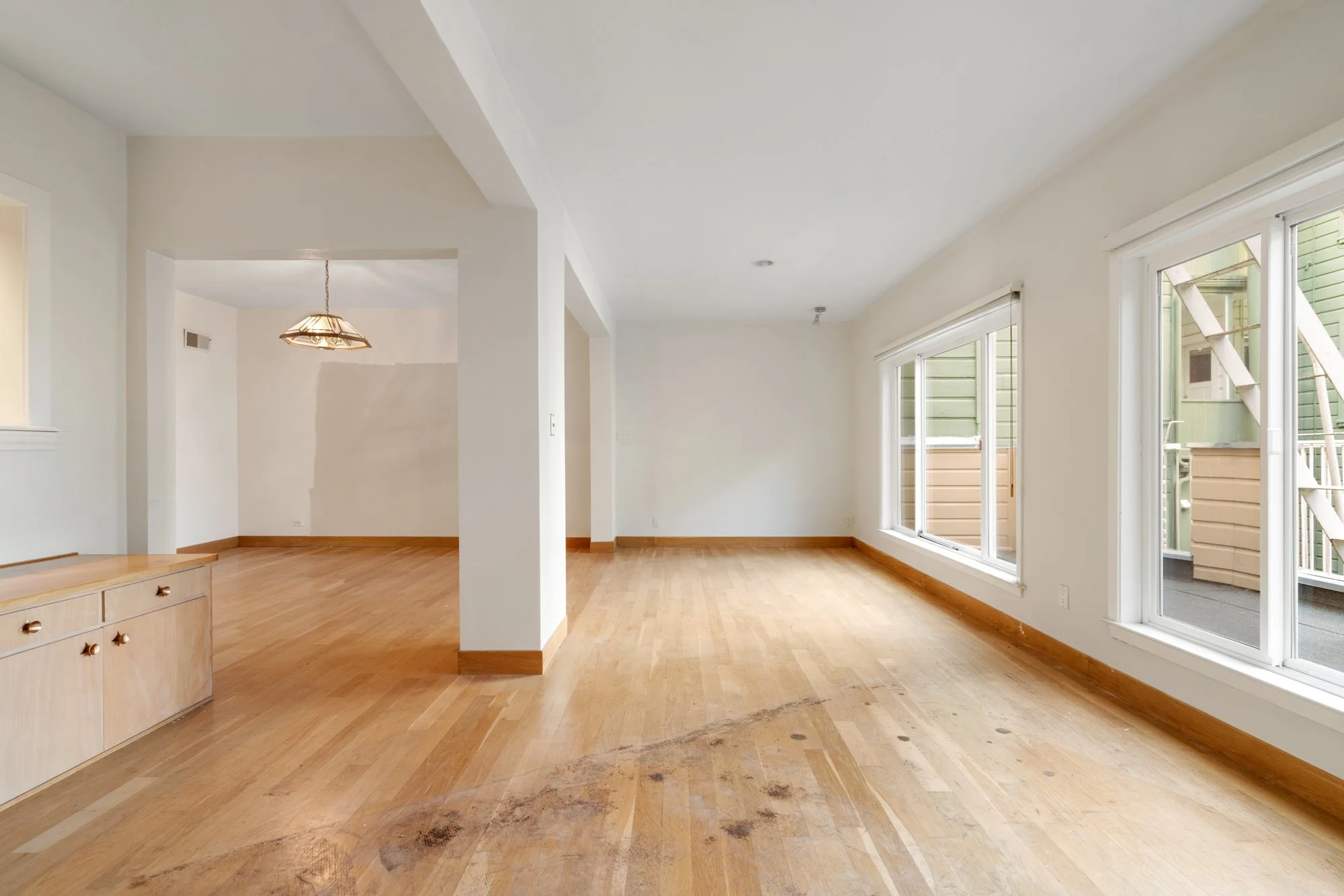 Empty living room with large windows, hardwood floors, and white walls, showing black stains on the floor.