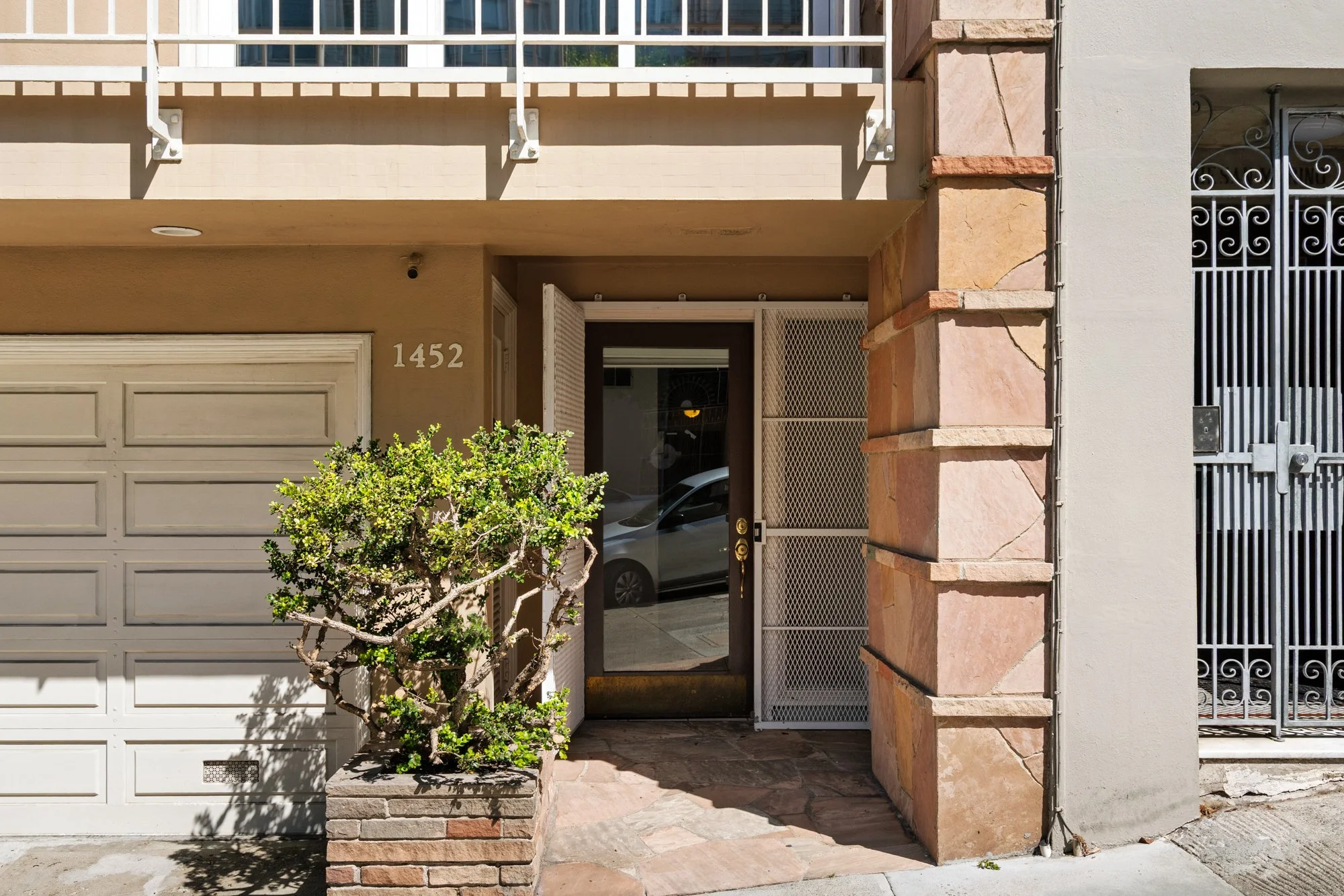 The front entrance of a beige and stone residential building showing a garage door on the left, a door with a screen door in the center, a small tree in front, and a gated area on the right.