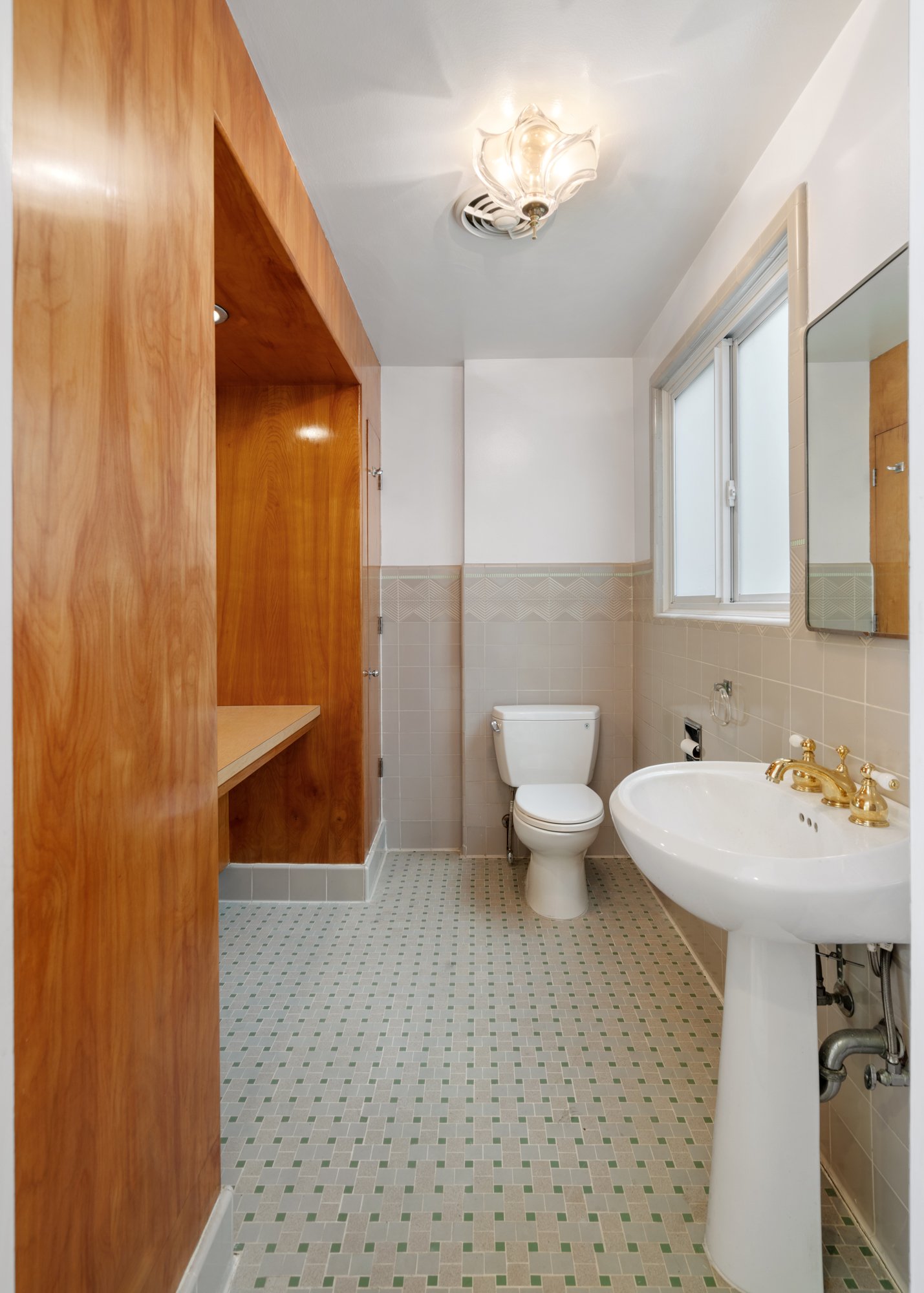 Interior of a small bathroom with a wooden wall on the left, a white toilet, white sink with gold fixtures, a mirror, a window, and a tiled floor and wall