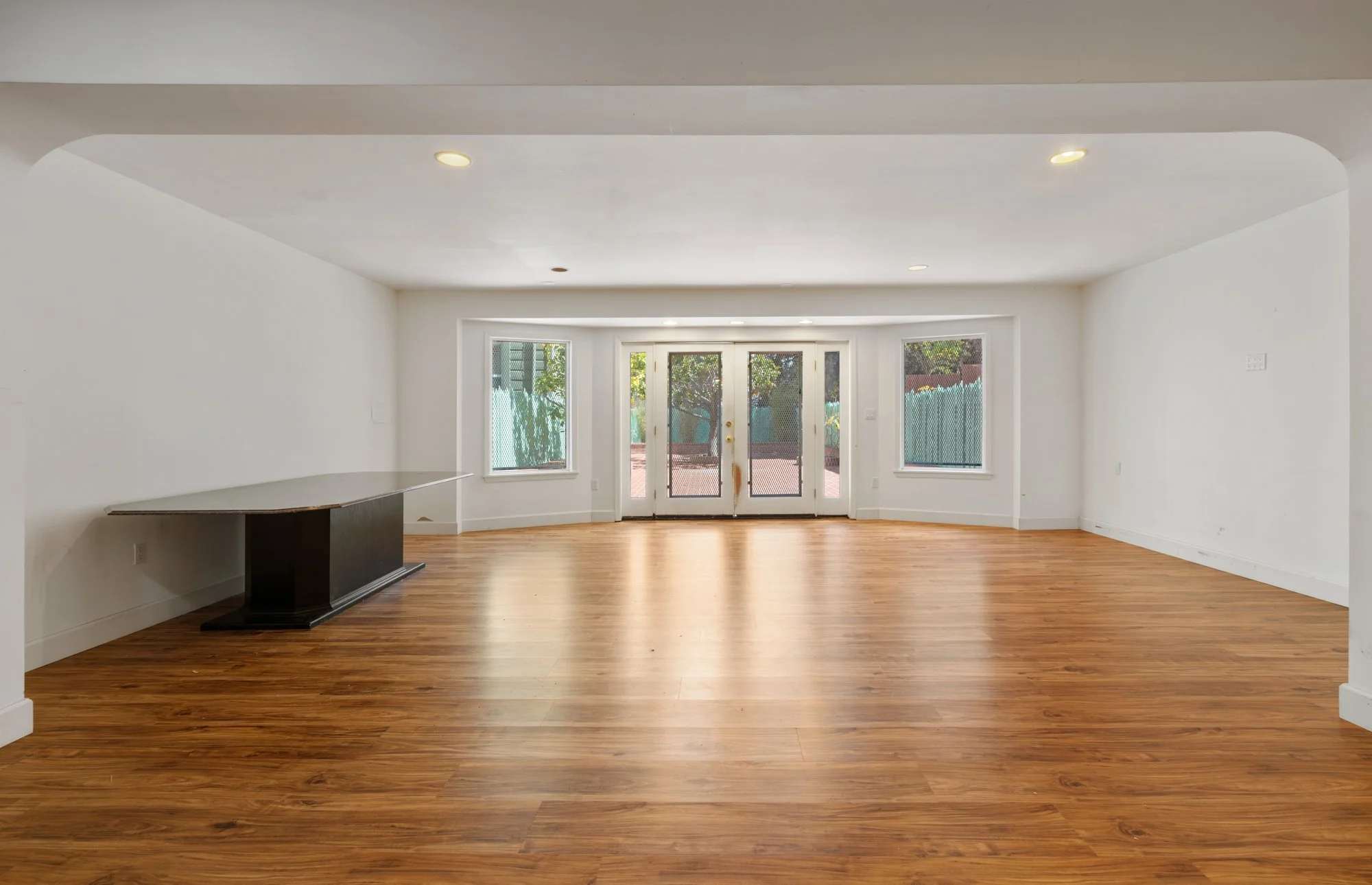 Empty living room with hardwood floors, white walls, and large windows with a door leading outside.