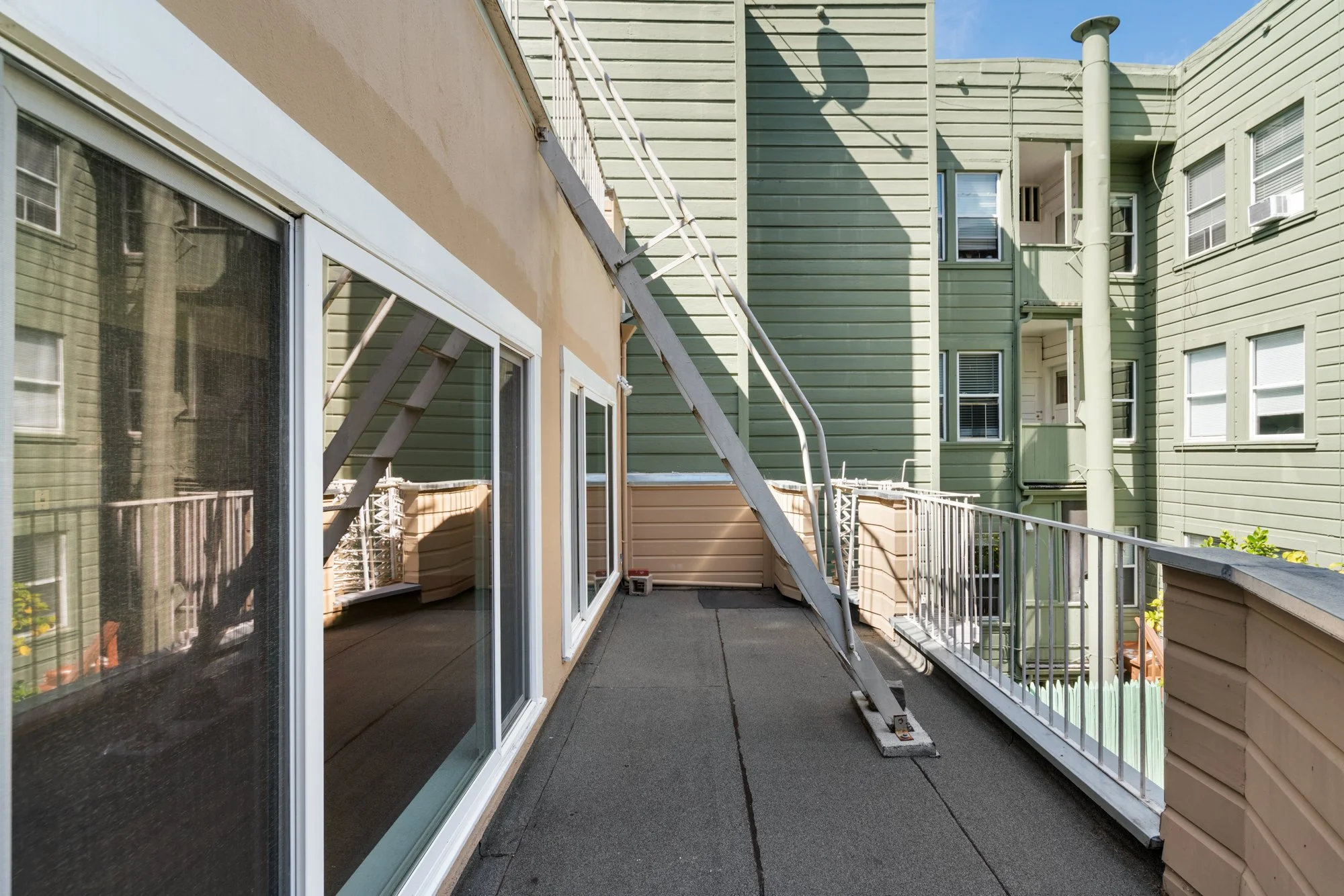 Apartment balcony with sliding glass door, railing, and green and beige building exterior, shadow of a lamp on the wall, and a ladder leaning against the wall.