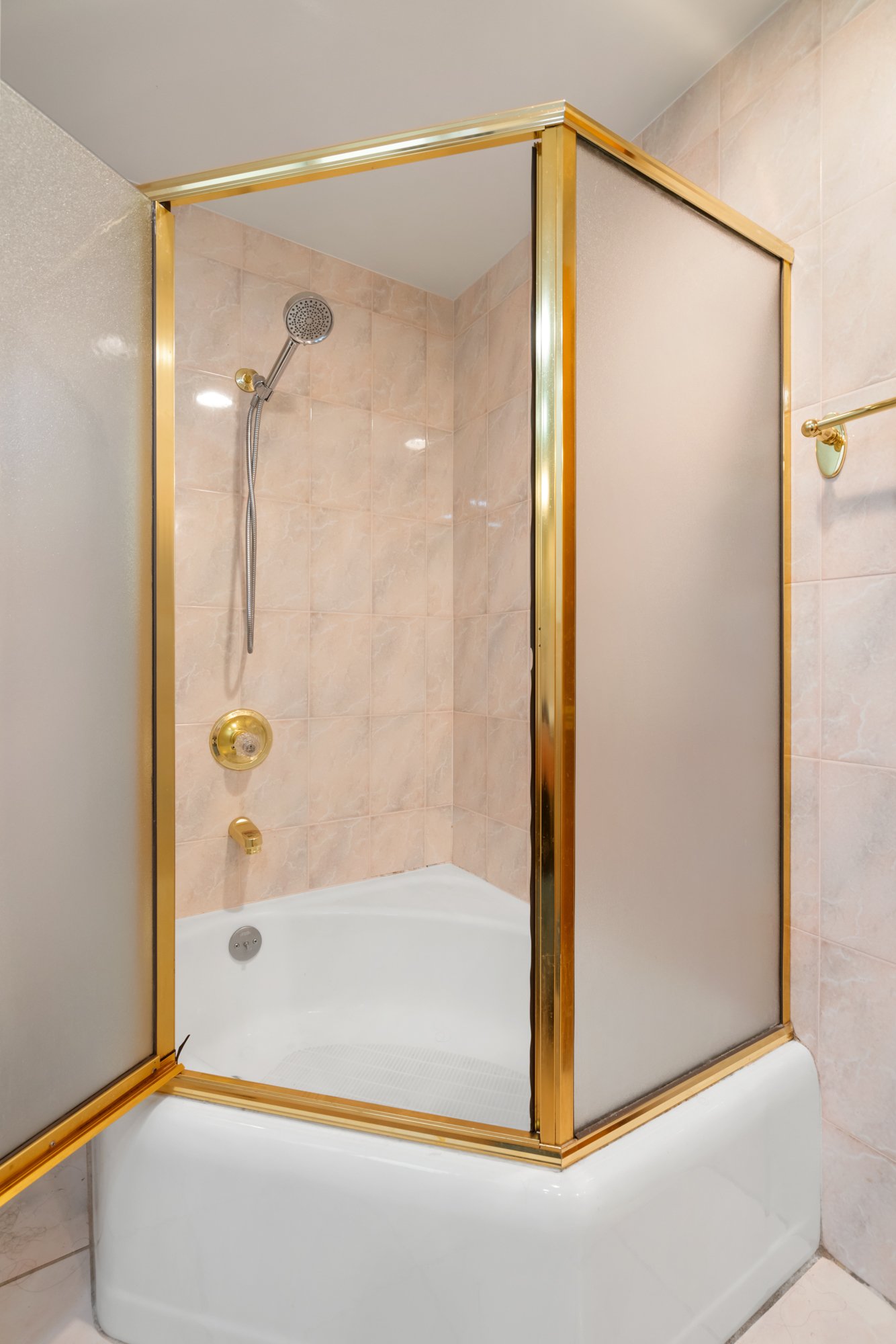 A bathtub with a glass shower enclosure featuring a gold trim, with beige tiled walls and a handheld showerhead.