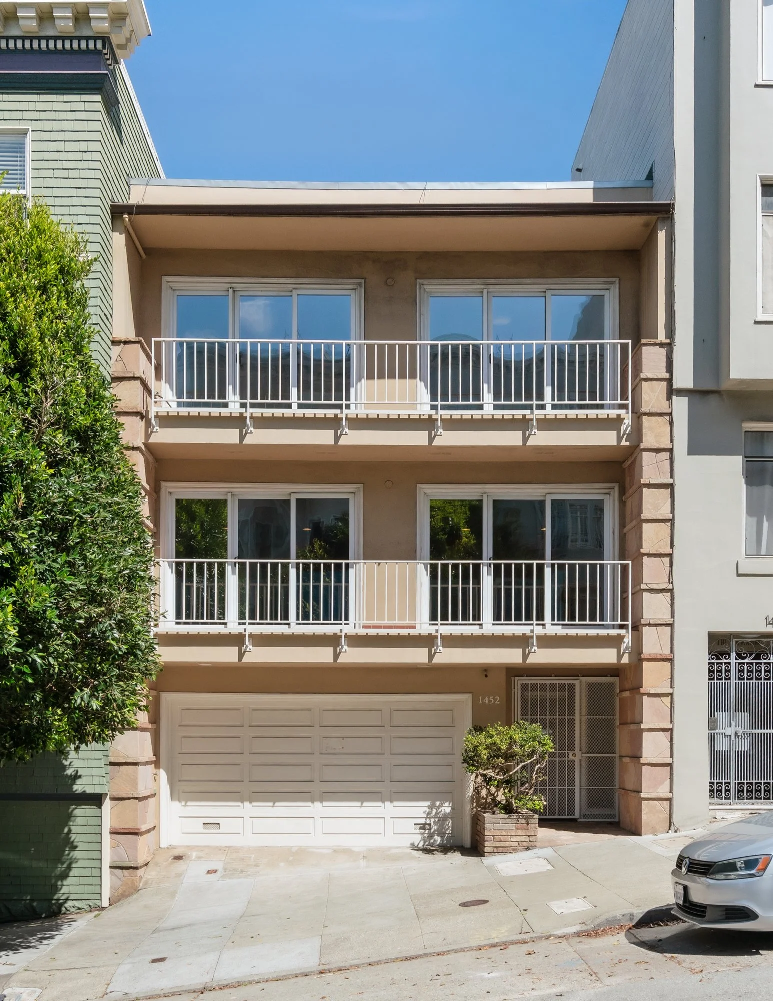 Three-story residential building with beige and green facades, white balconies, garage door, and a sidewalk in front. A silver car is parked on the street to the right. Clear blue sky overhead.