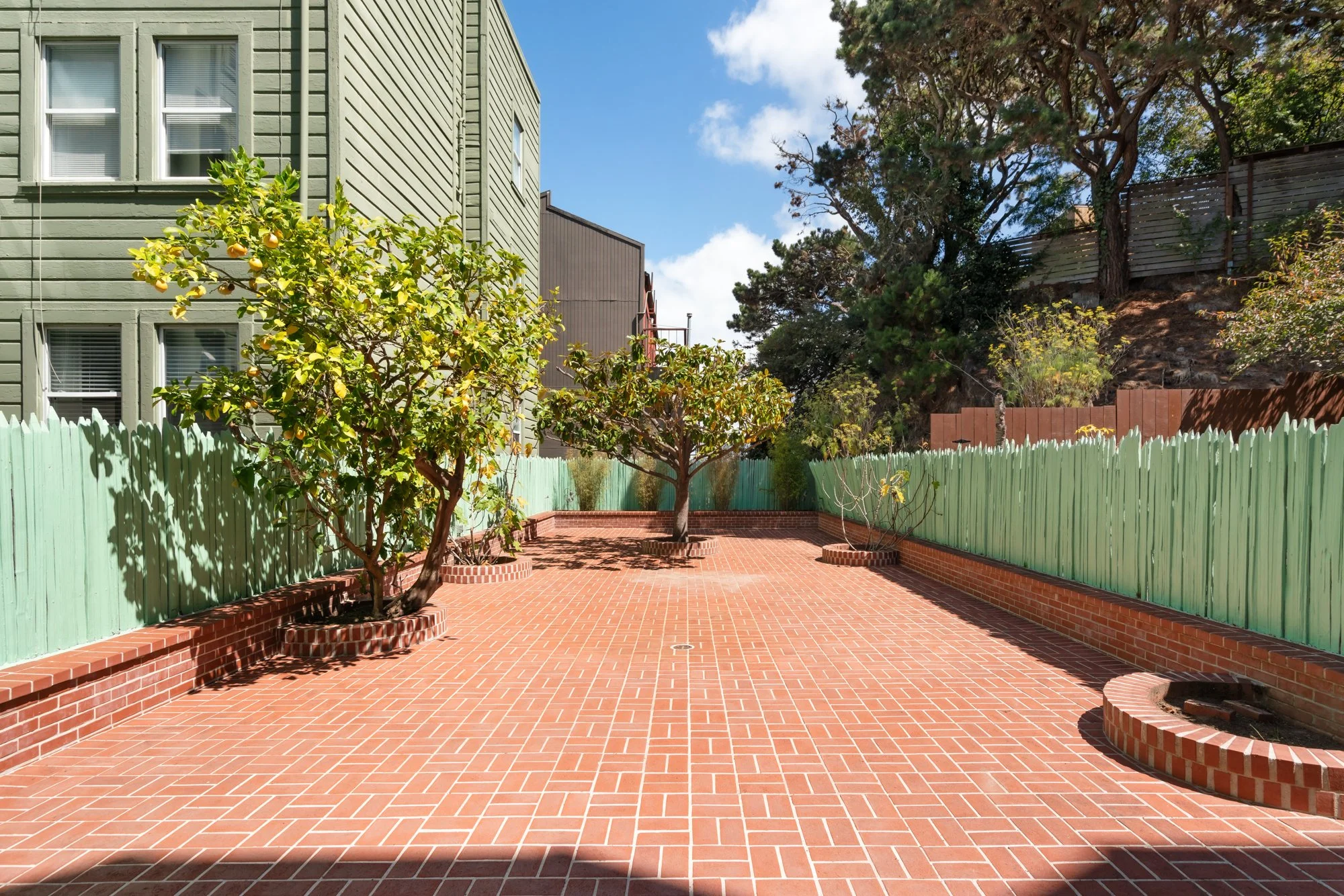 A patio with red brick paving, several small lemon trees in brick planters, a green wooden fence, and a house with beige siding in the background. Some trees and a wooden fence are visible on a hill in the background, with a blue sky and few clouds o