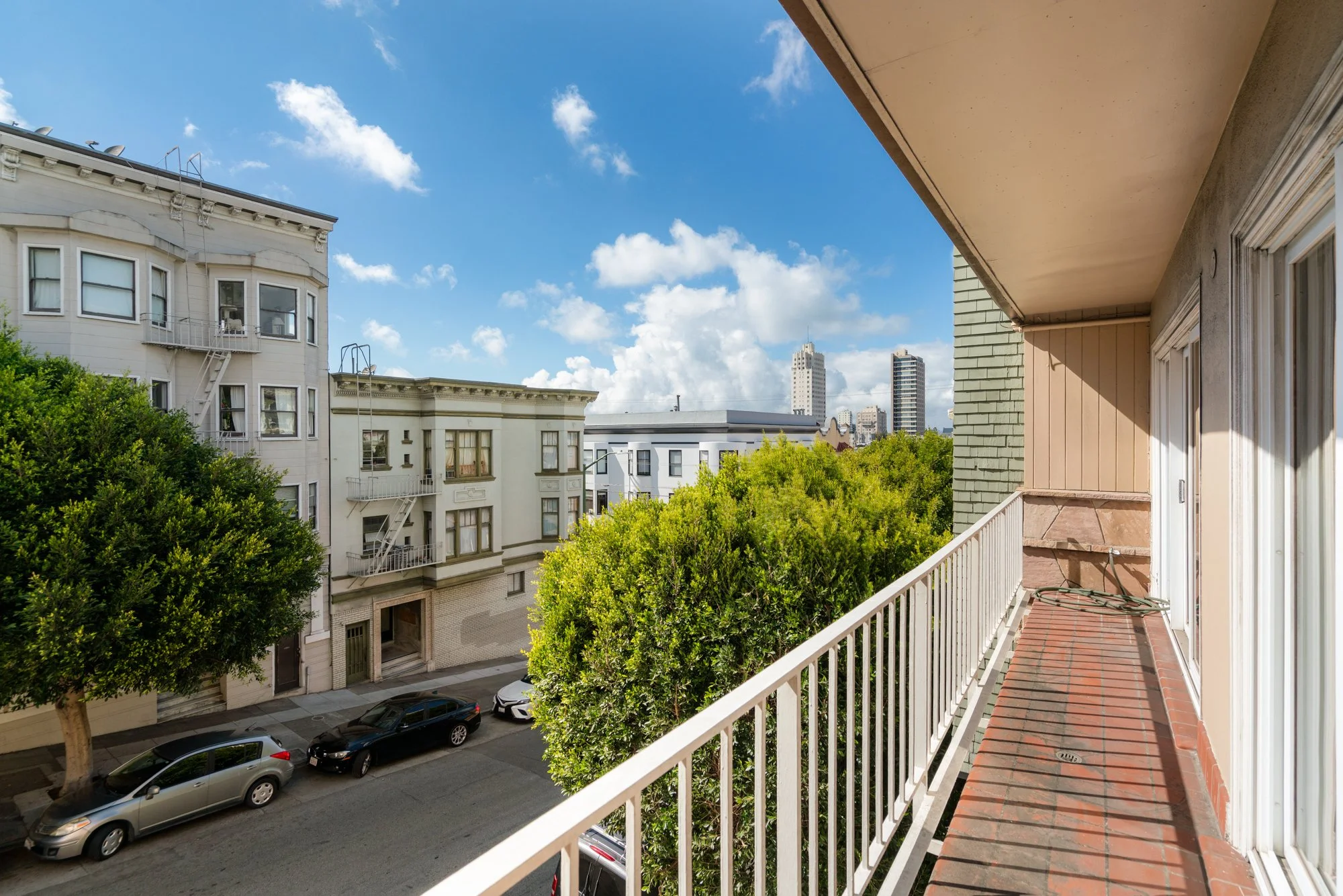 View from a balcony showing a city street with parked cars and trees, neighboring buildings, and a clear blue sky with scattered clouds.