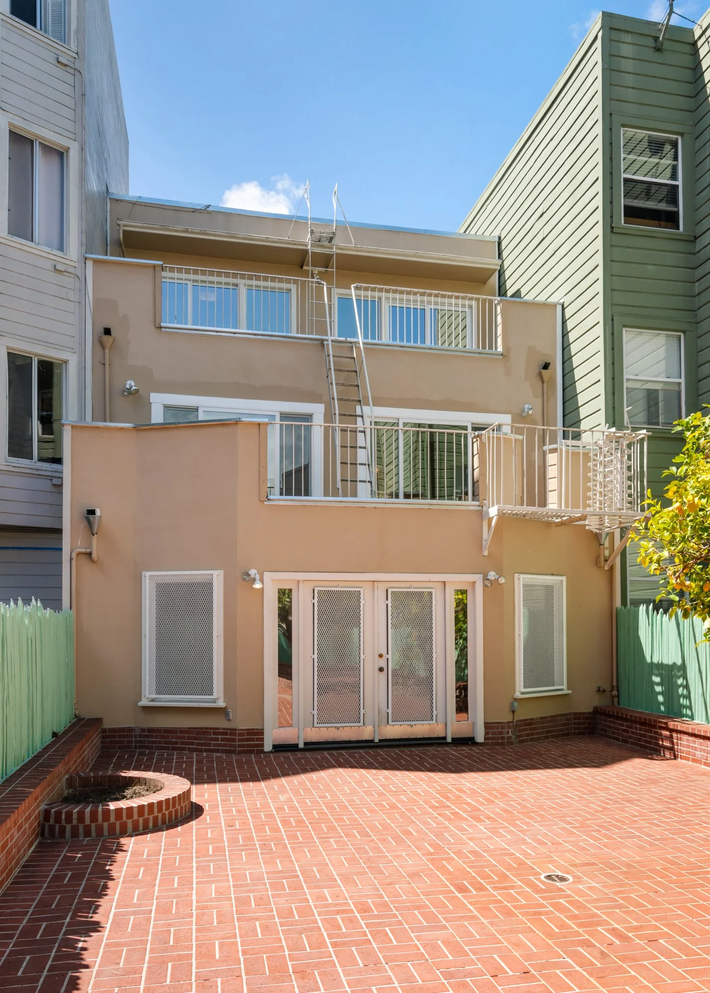 Backyard with red brick patio, green wooden fences, and beige residential building with balconies and white doors.