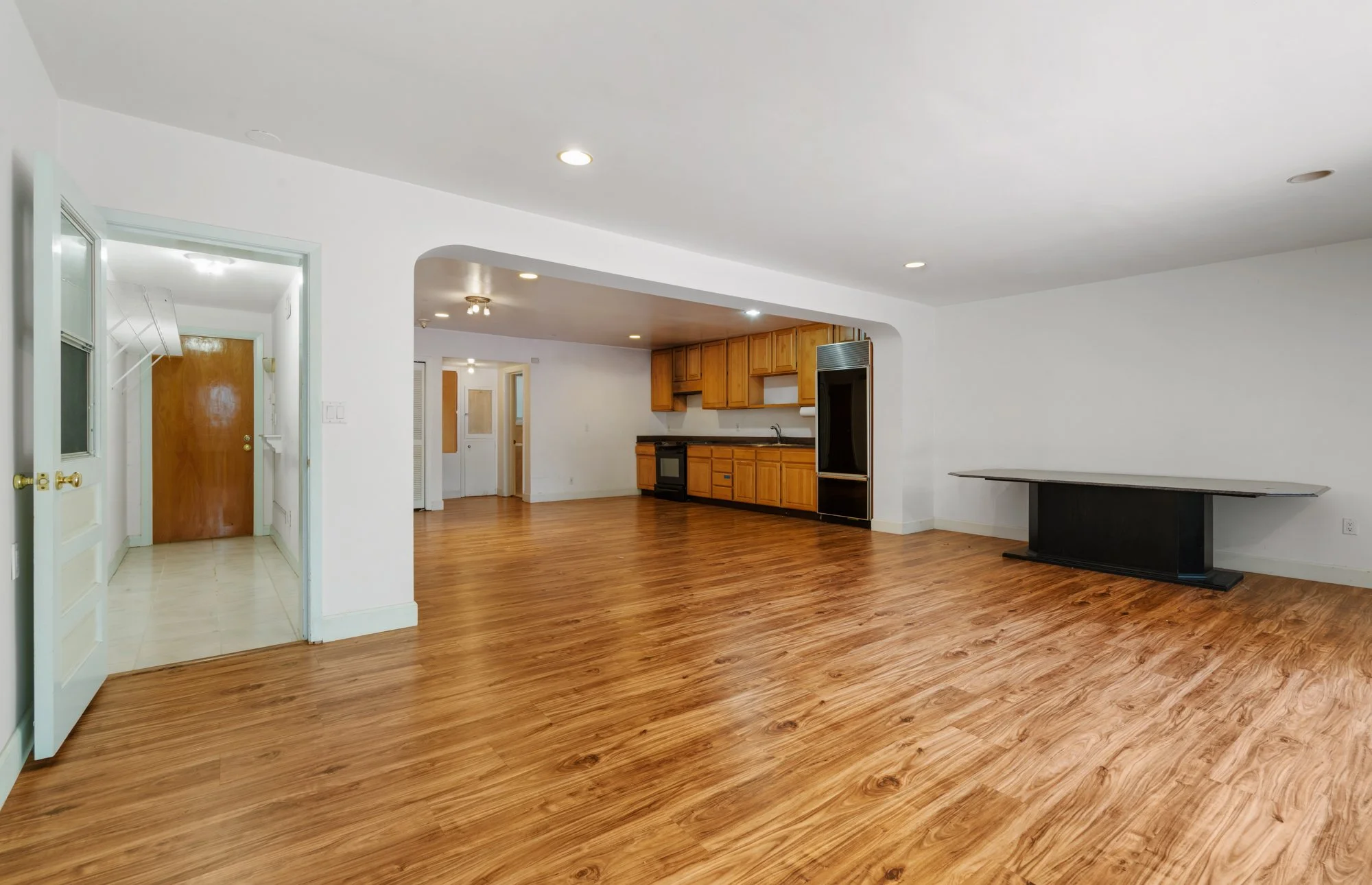 Open-concept living space with hardwood floors, white walls, ceiling lights, a kitchen with wooden cabinets, a stove, and a refrigerator, and a large table or desk against the wall.