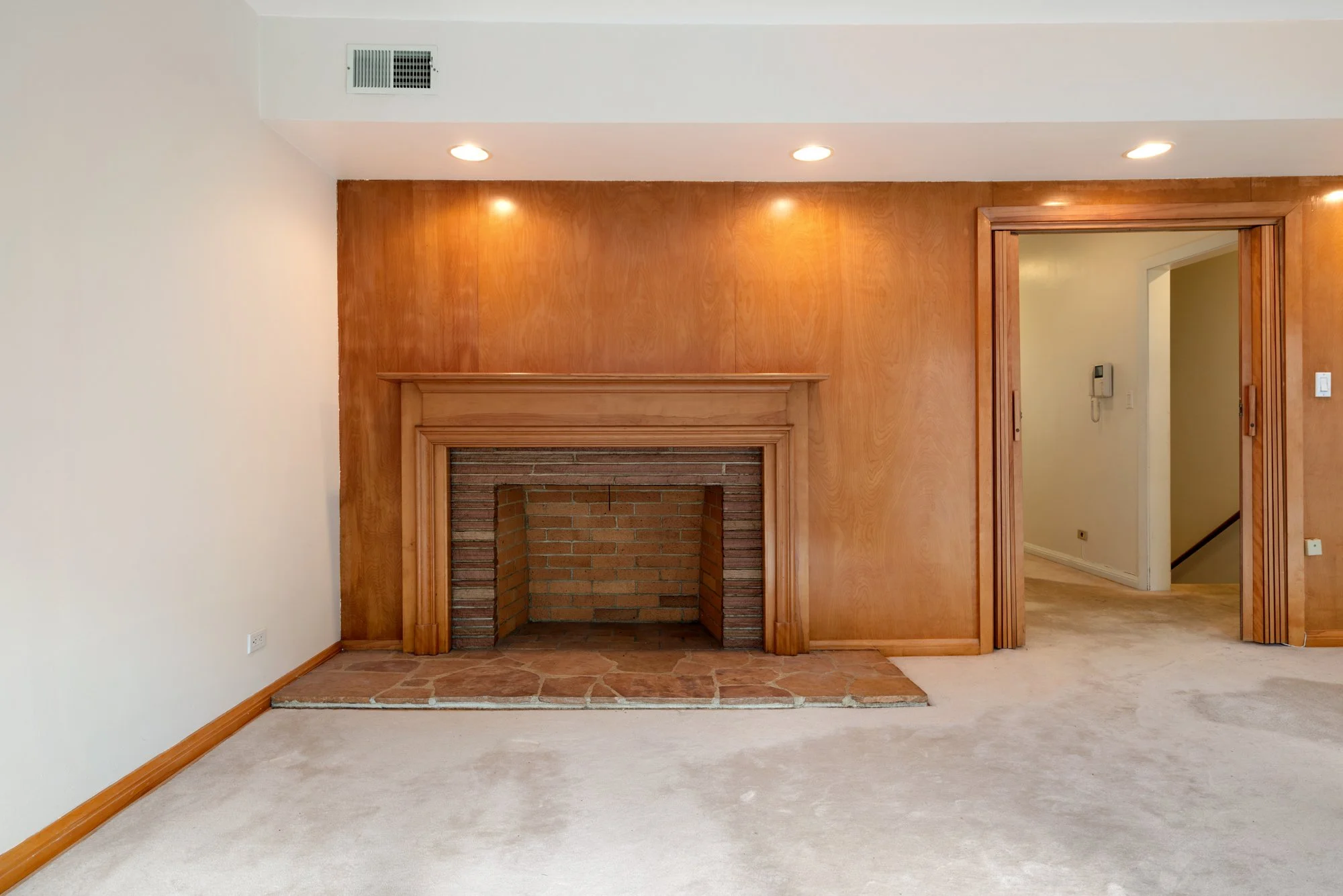 Living room with a brick fireplace, wooden mantel, wooden wall, and a doorway leading to a hall with a beige wall and phone