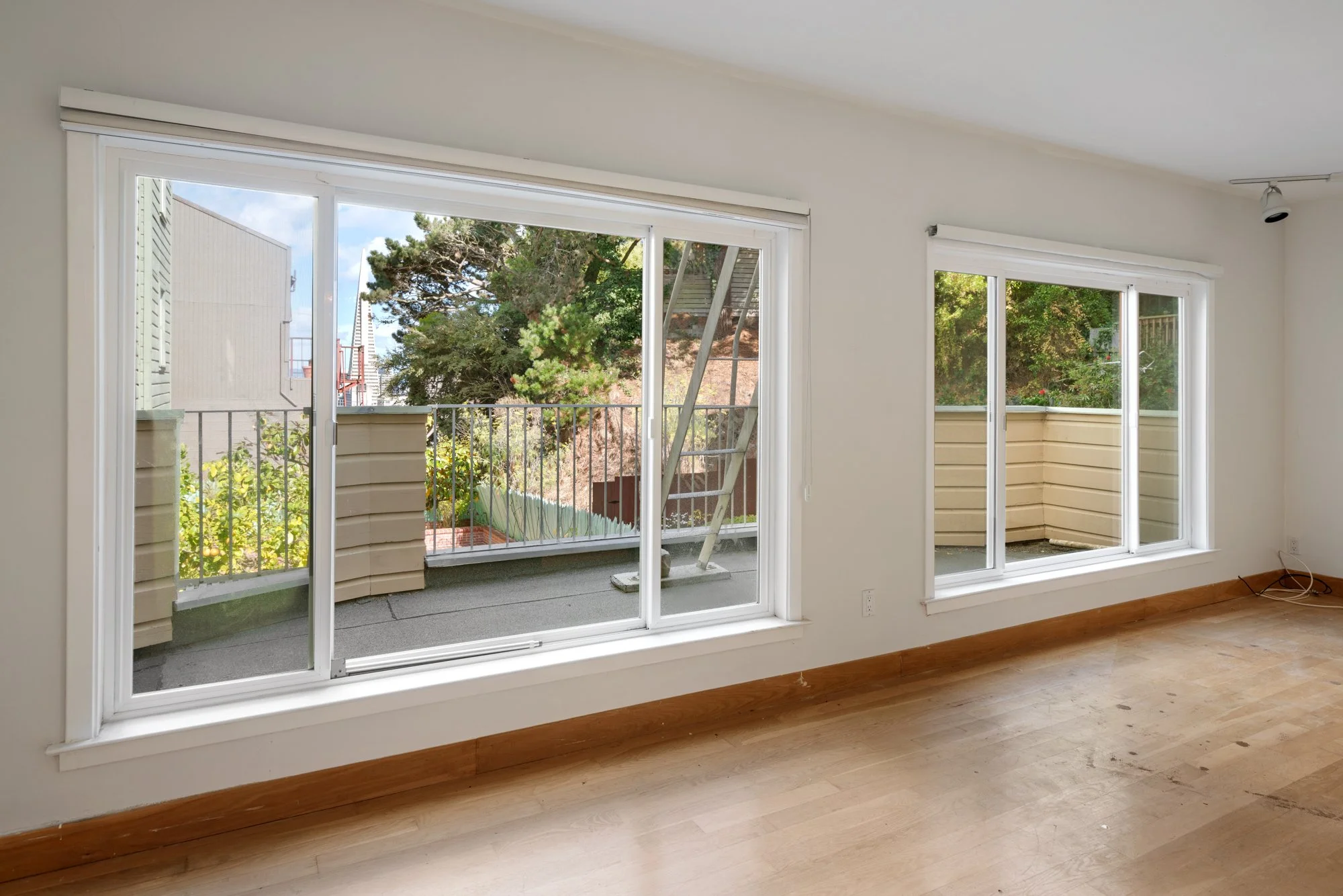 Empty room with hardwood floors and large sliding glass doors opening to a balcony with a metal railing and surrounding trees.