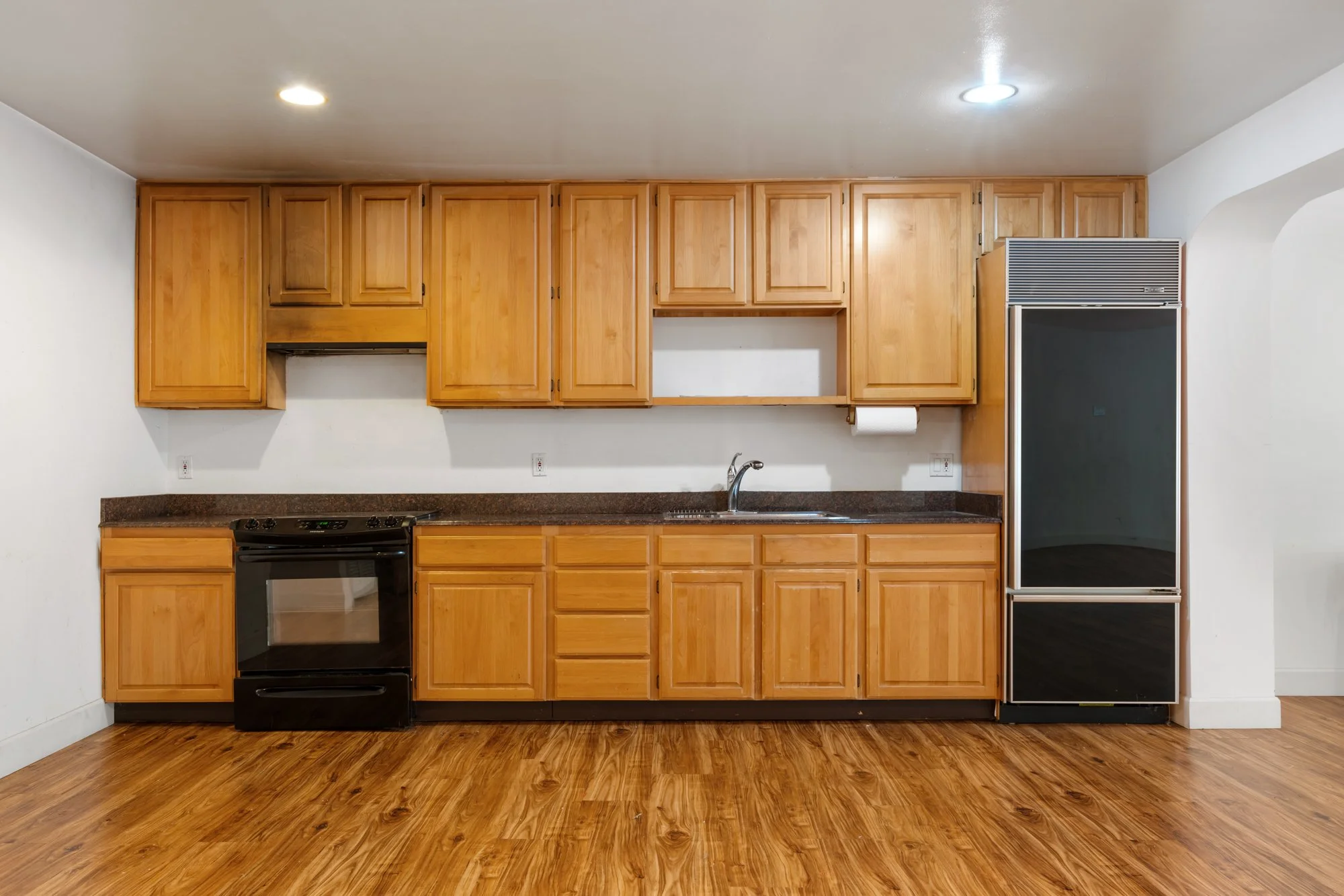 Kitchen with wooden cabinets, black stove, and a refrigerator, with hardwood flooring and white walls.