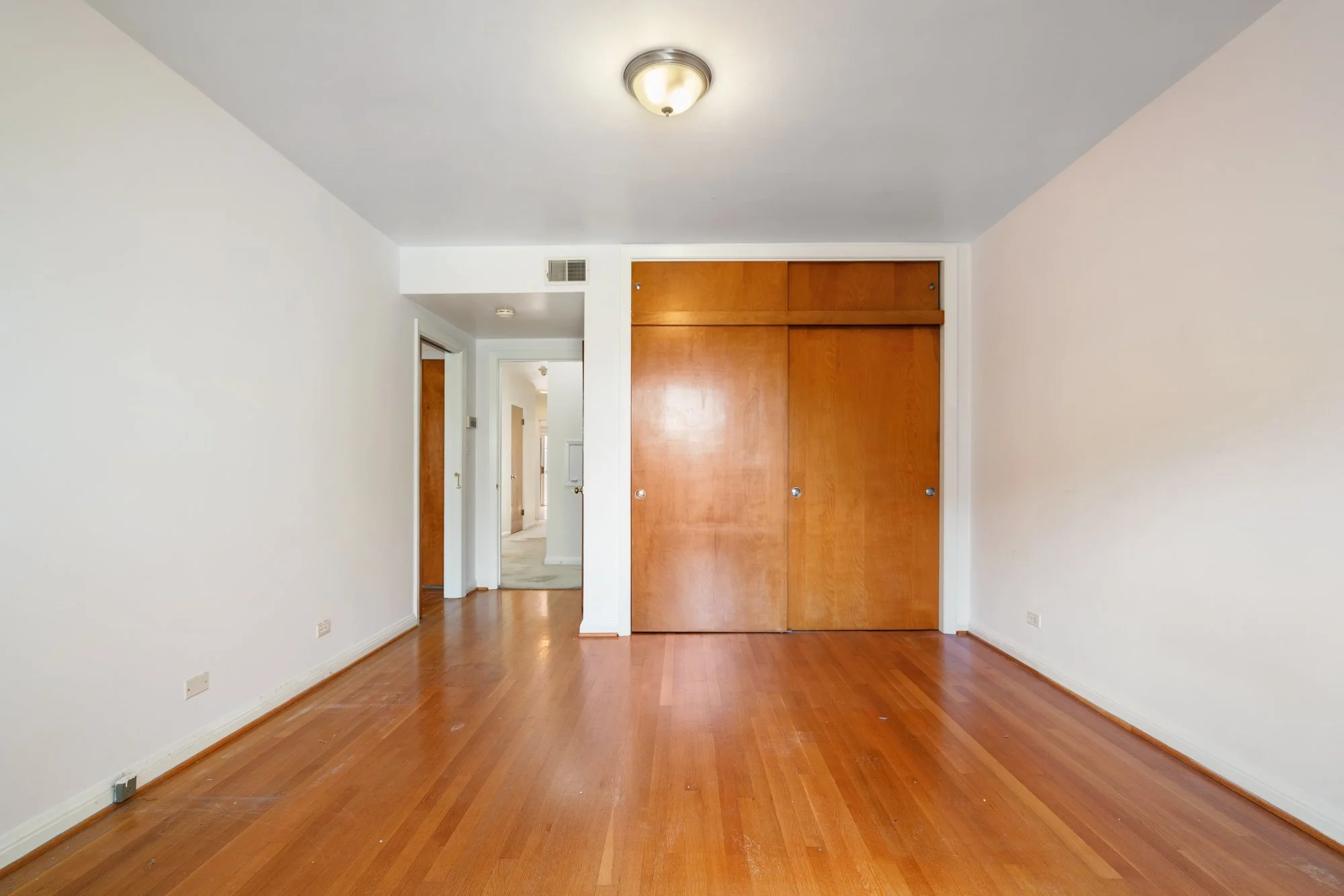 Empty room with wooden floor, white walls, sliding closet doors, and hallway with doors leading to other rooms.