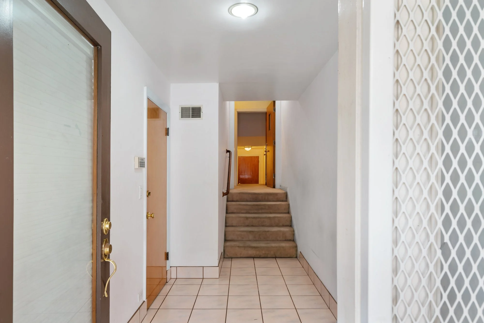 Entryway with tiled floor, wooden and glass doors, staircase with beige carpet, white walls, ceiling light, and wall-mounted thermostat.