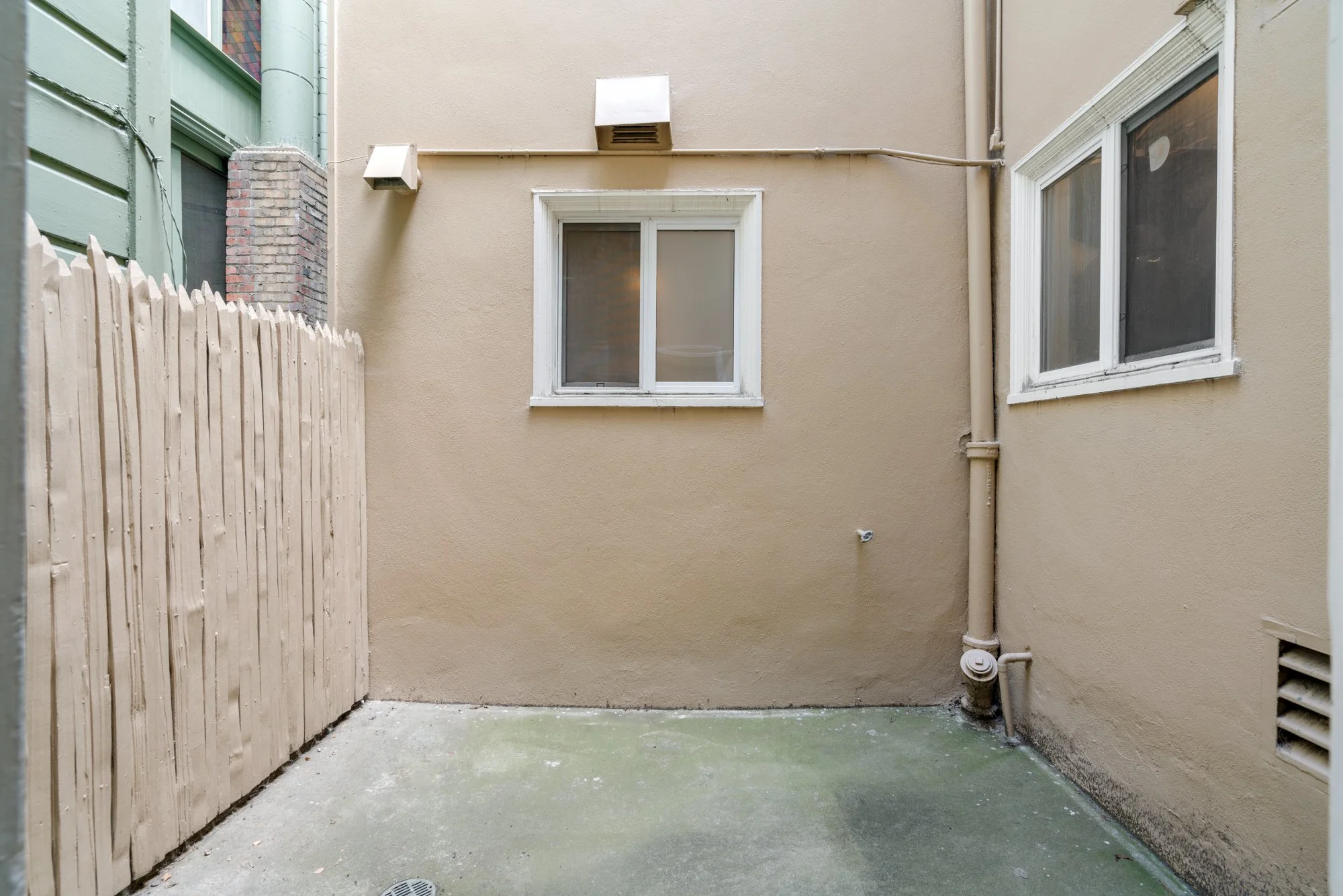 An empty outdoor backyard with concrete flooring, beige walls, a wooden fence on the left, and two windows on the right wall, along with some utility pipes and vents.