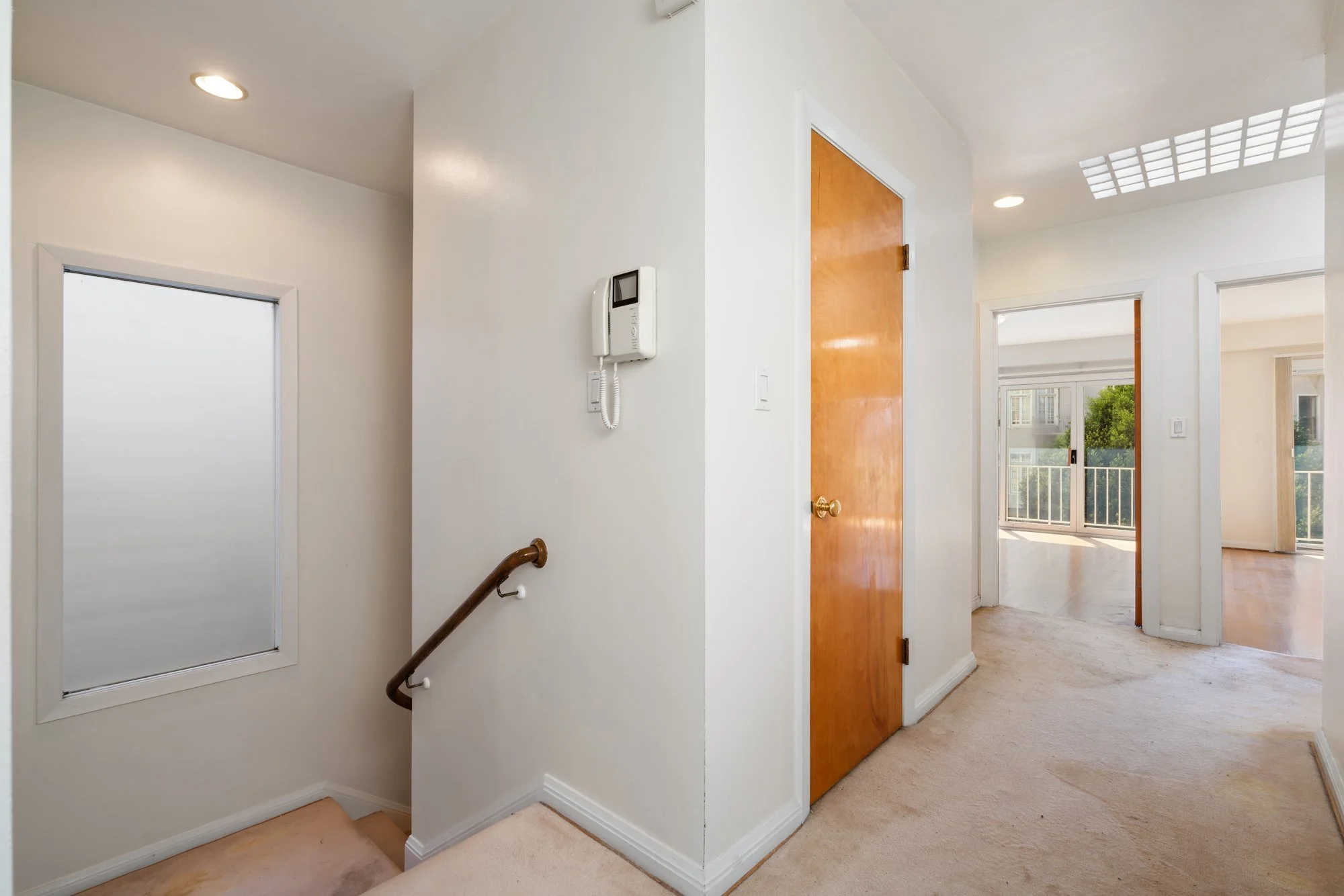 Interior view of a residential space showing a hallway with a wooden door, a white wall with a wall-mounted intercom, and large windows letting in natural light.