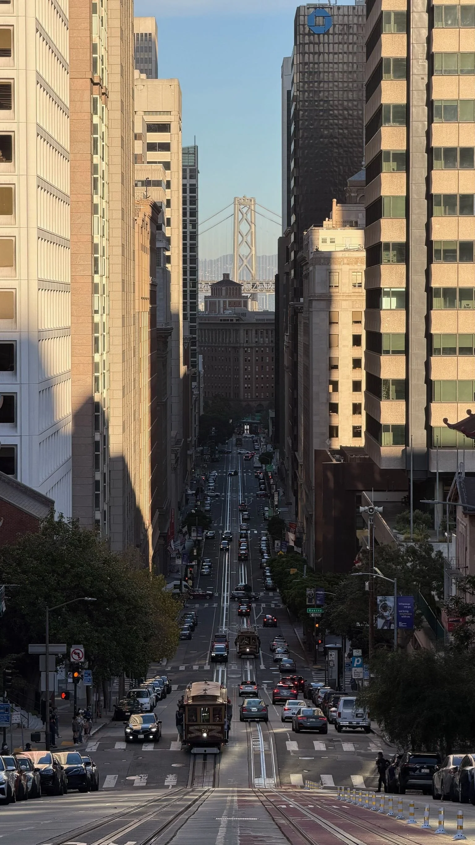 A city street viewed from a hill, with cars and a cable-driven streetcar in the foreground, flanked by tall office buildings on both sides, leading towards the San Francisco Bay Bridge in the distance.