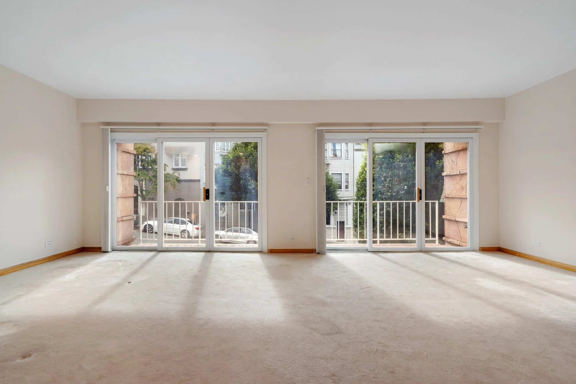 Empty living room with large sliding glass doors leading to a balcony, sunlight streaming in, and a view of neighboring buildings and trees.