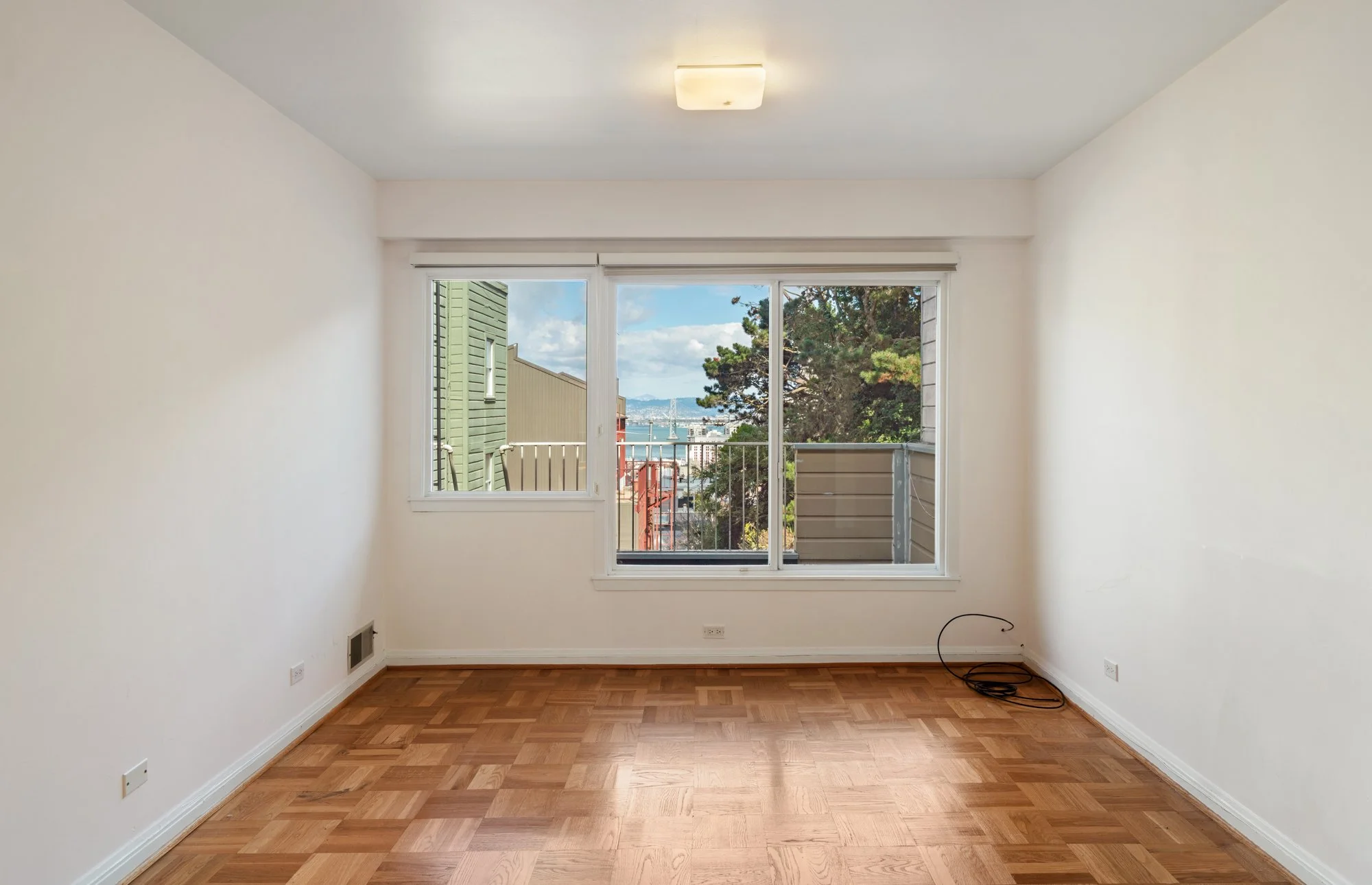Empty living room with hardwood floors, white walls, a large window showing trees, buildings, and a cloudy sky outside, and a ceiling light fixture.