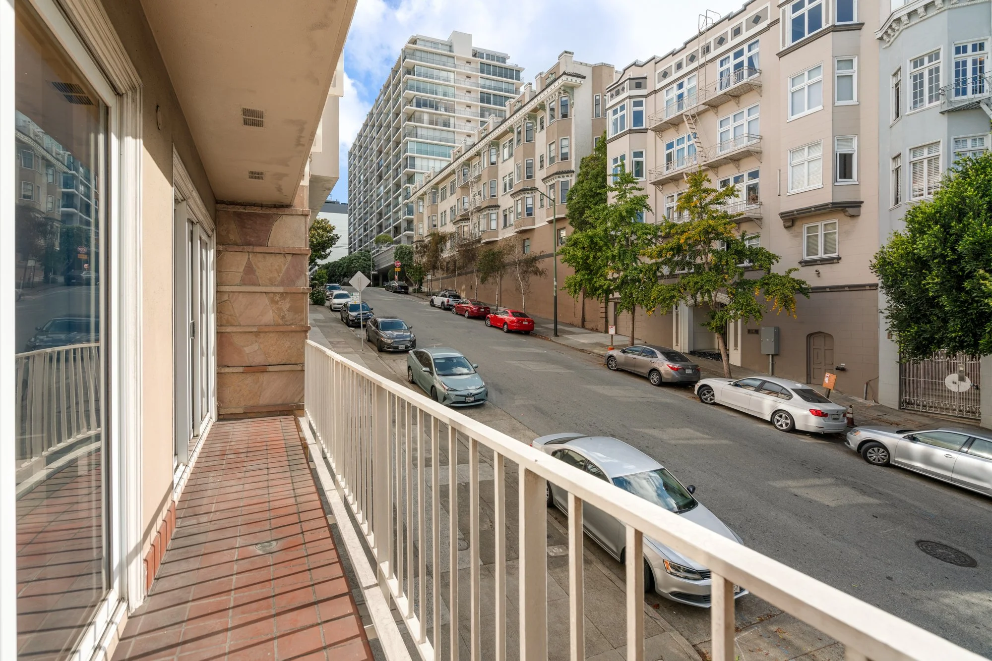 View from a balcony showing a street lined with parked cars and multi-story residential buildings under a partly cloudy sky.