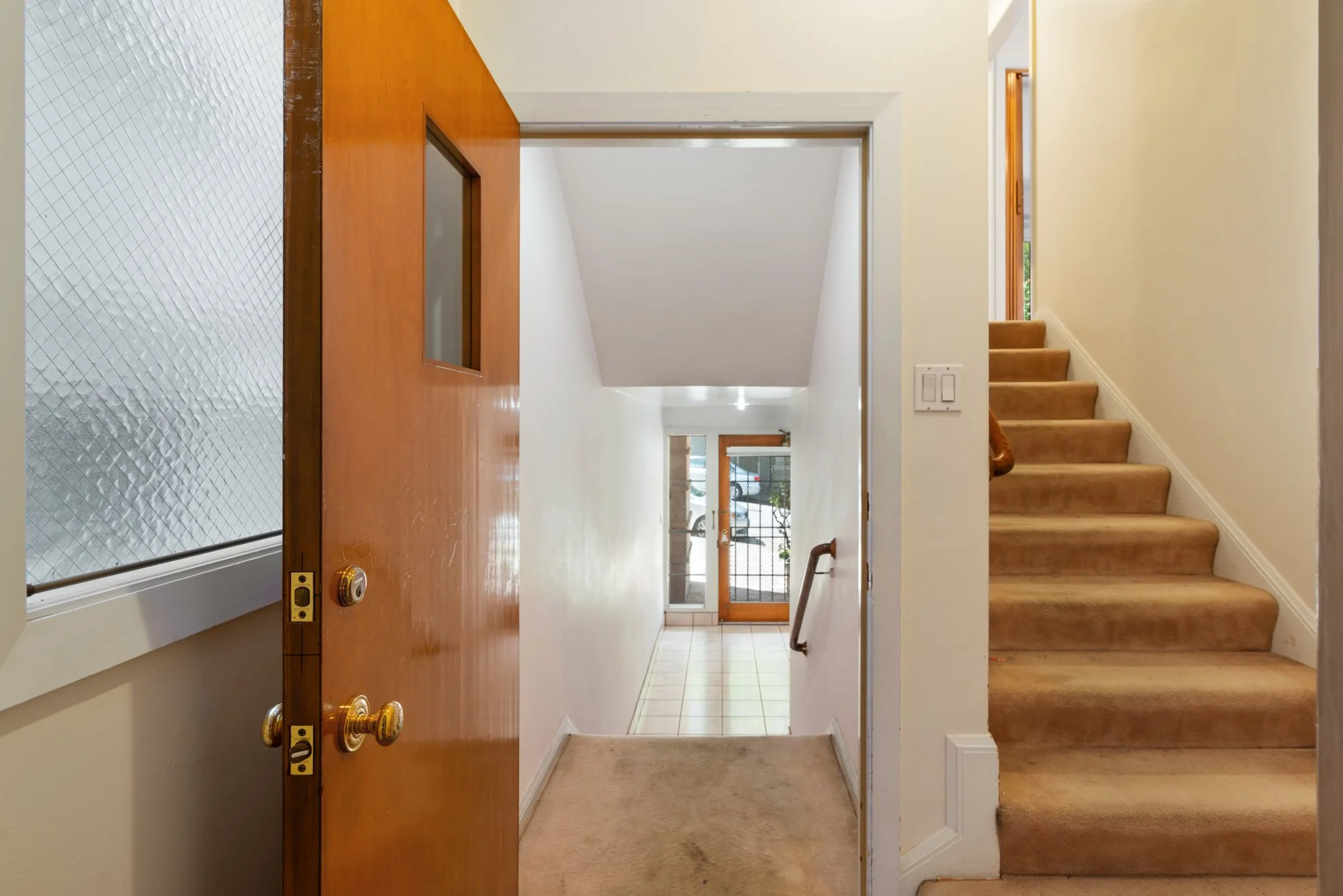 Interior entryway with stairs on the right, glass door at the far end, and wooden accents including a doorframe and a door with a glass panel. The walls are painted light, and the floor has beige carpet transitioning to tiled flooring near the entran