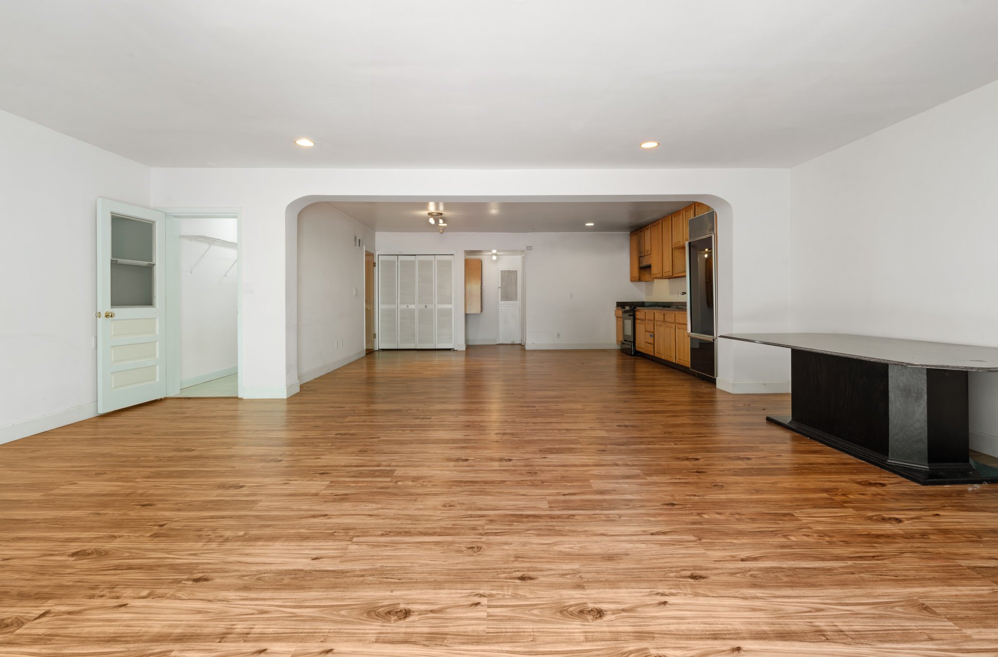 Empty living room with wooden floors, white walls, and recessed ceiling lights, connected to a kitchen area with wooden cabinets and a black refrigerator.