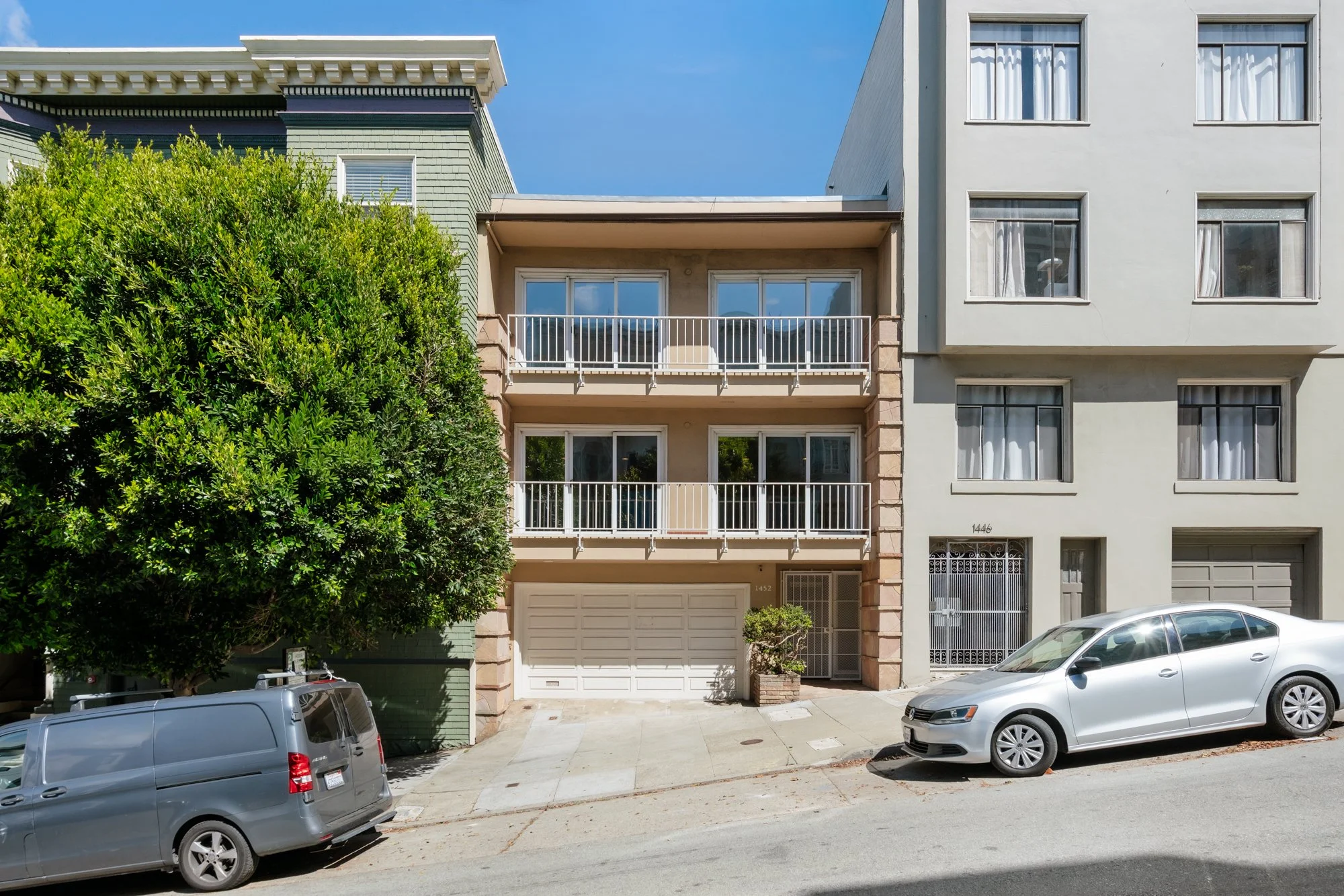 A modern residential building with a green facade on the left and a white facade on the right, featuring multiple balconies with white railings and ground-level garage doors. Cars are parked in front, and a large tree partially blocks the view of the