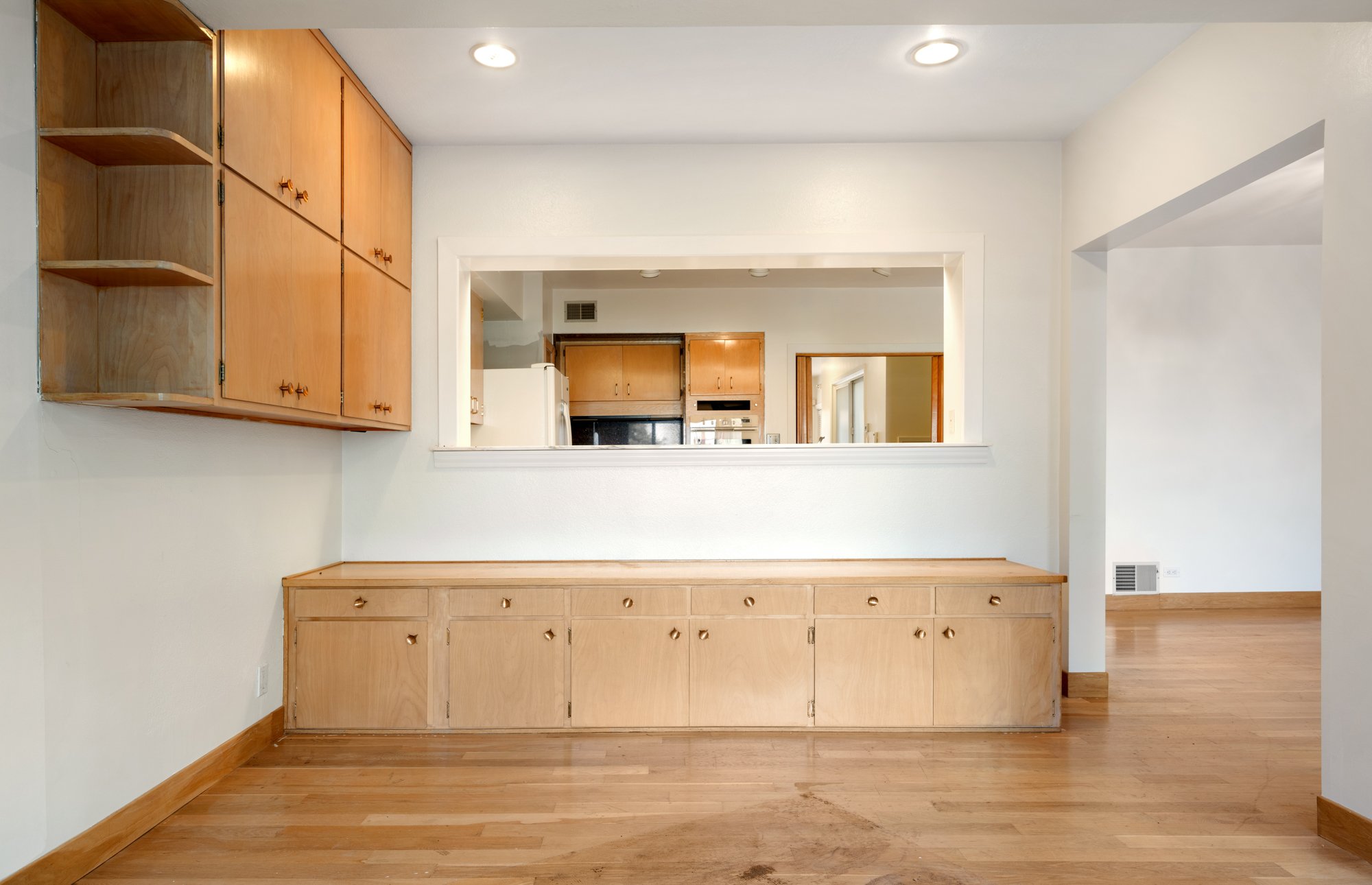 Interior view of a living room with wood flooring and built-in wooden cabinets. An open window-like partition frames the kitchen area with appliances visible in the background.