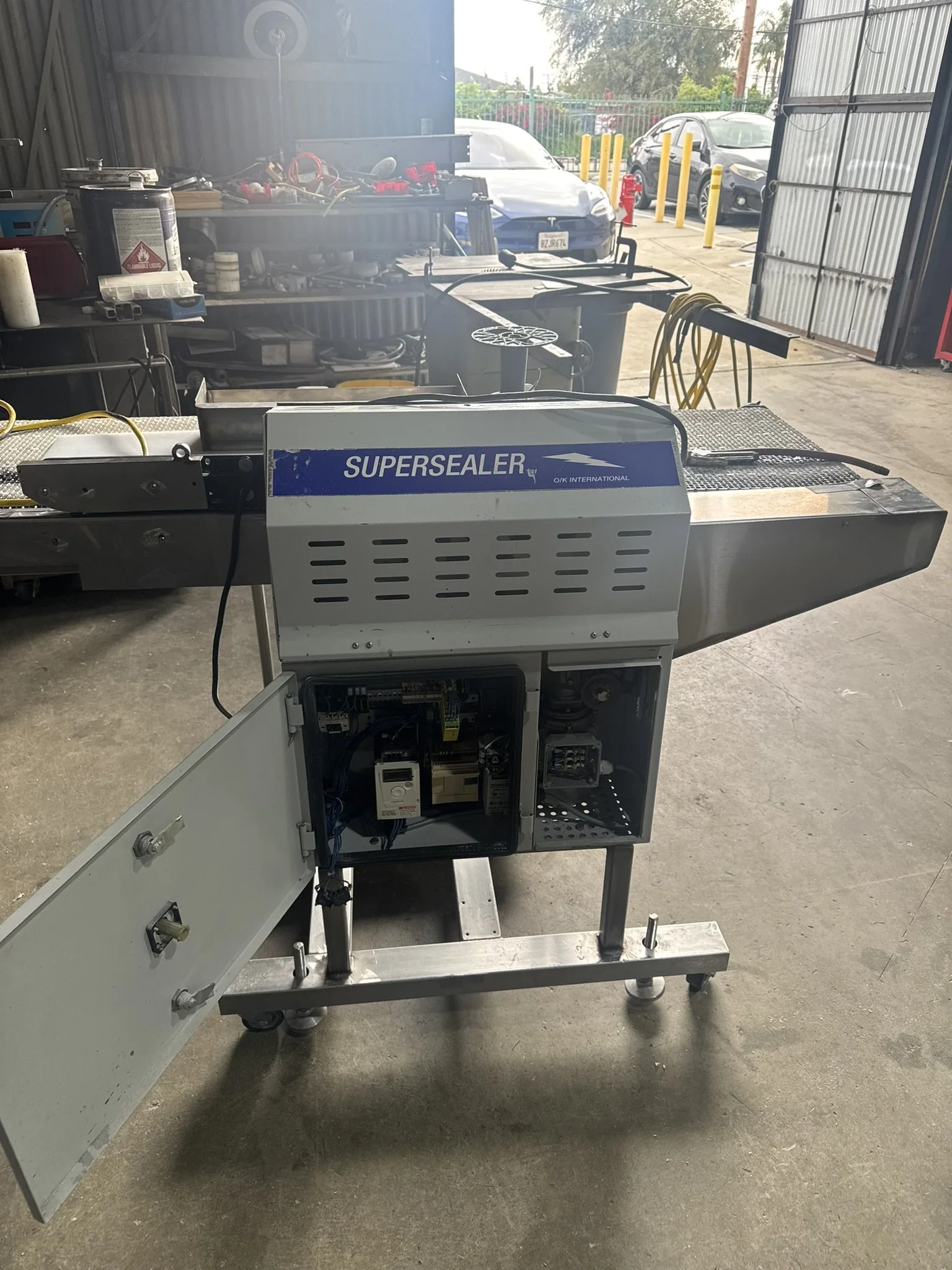 Metal worktable with open electrical control box and a conveyor oven labeled 'SUPERSEALER' in a workshop with cars outside in the background.