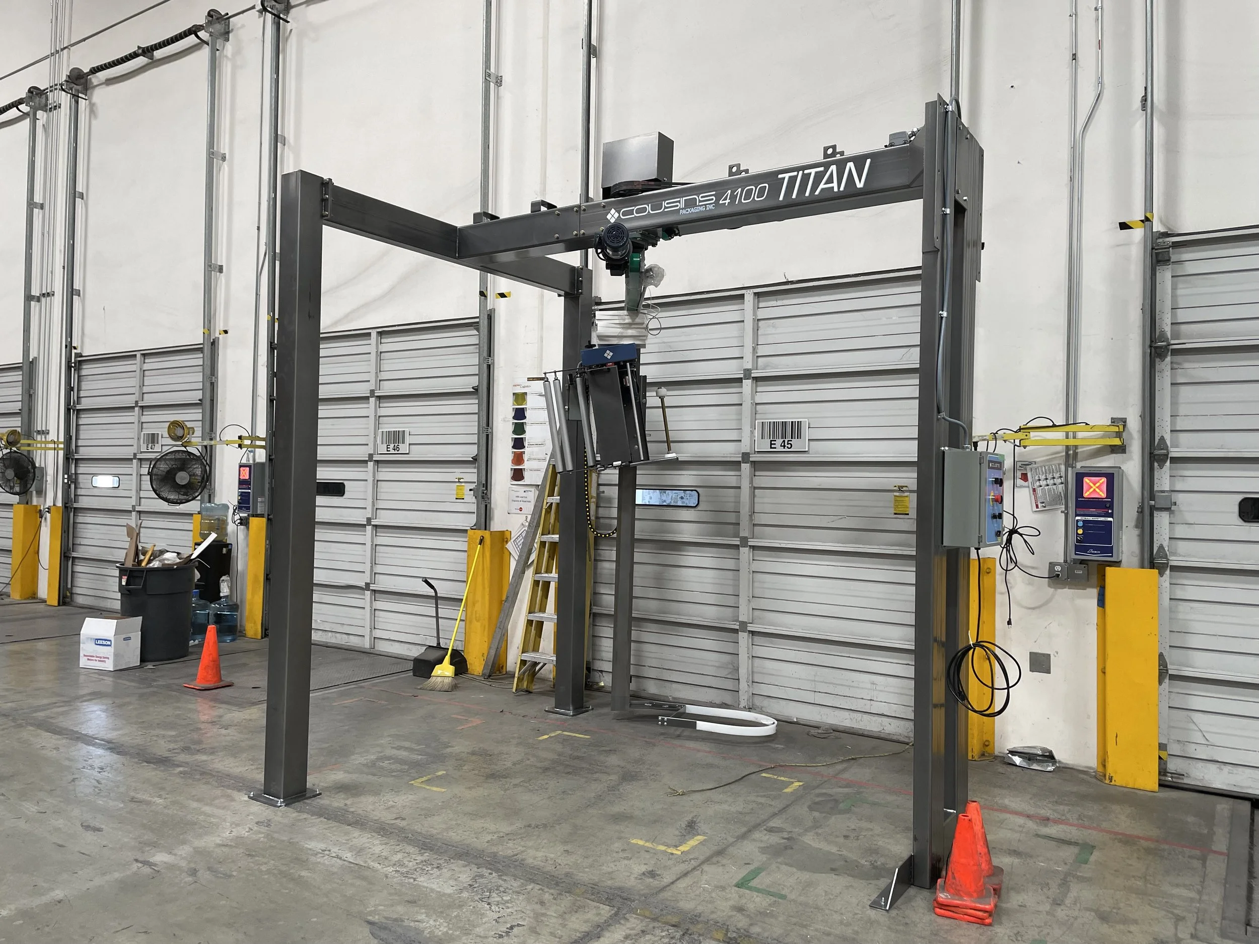 An industrial warehouse with a robotic arm machine labeled 'Cousins 4100 Titan', yellow safety barriers, orange cones, black fans, and various control panels.