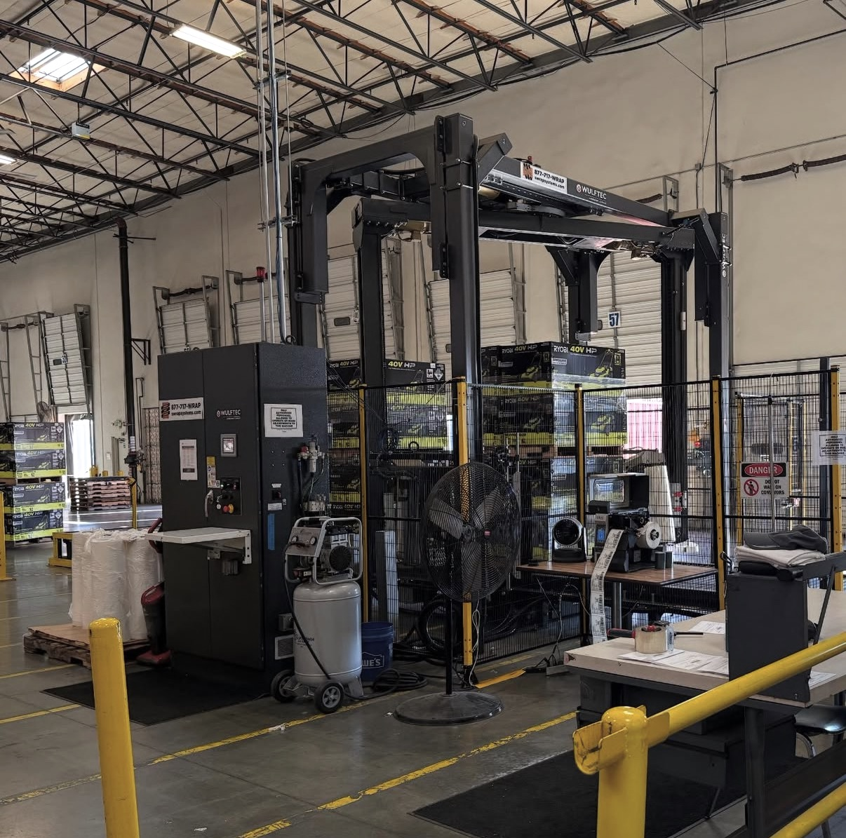 Industrial warehouse with machinery, safety fencing, and storage shelves containing boxes and equipment.