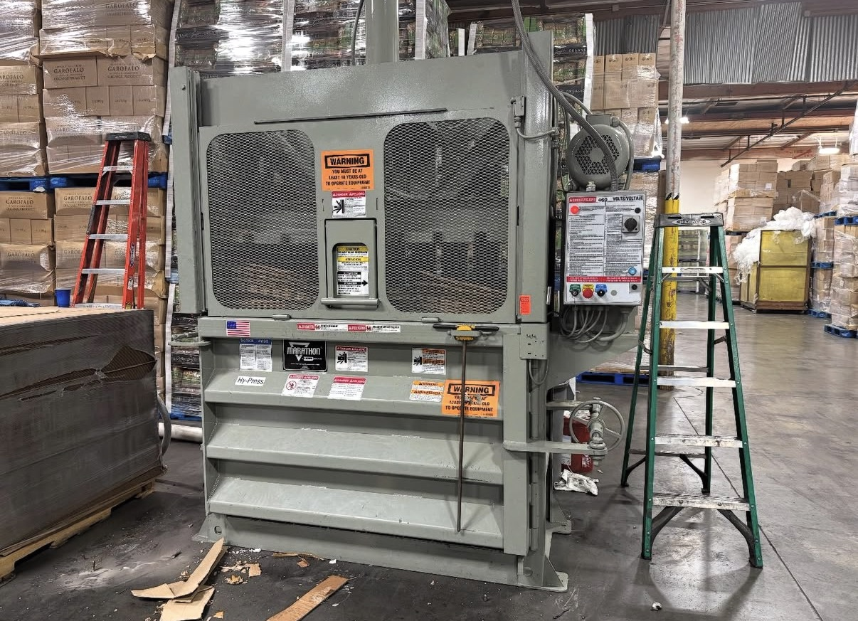 Industrial warehouse with large metal machine, four warning labels, a control panel, a green step ladder, and stacked boxes in the background.