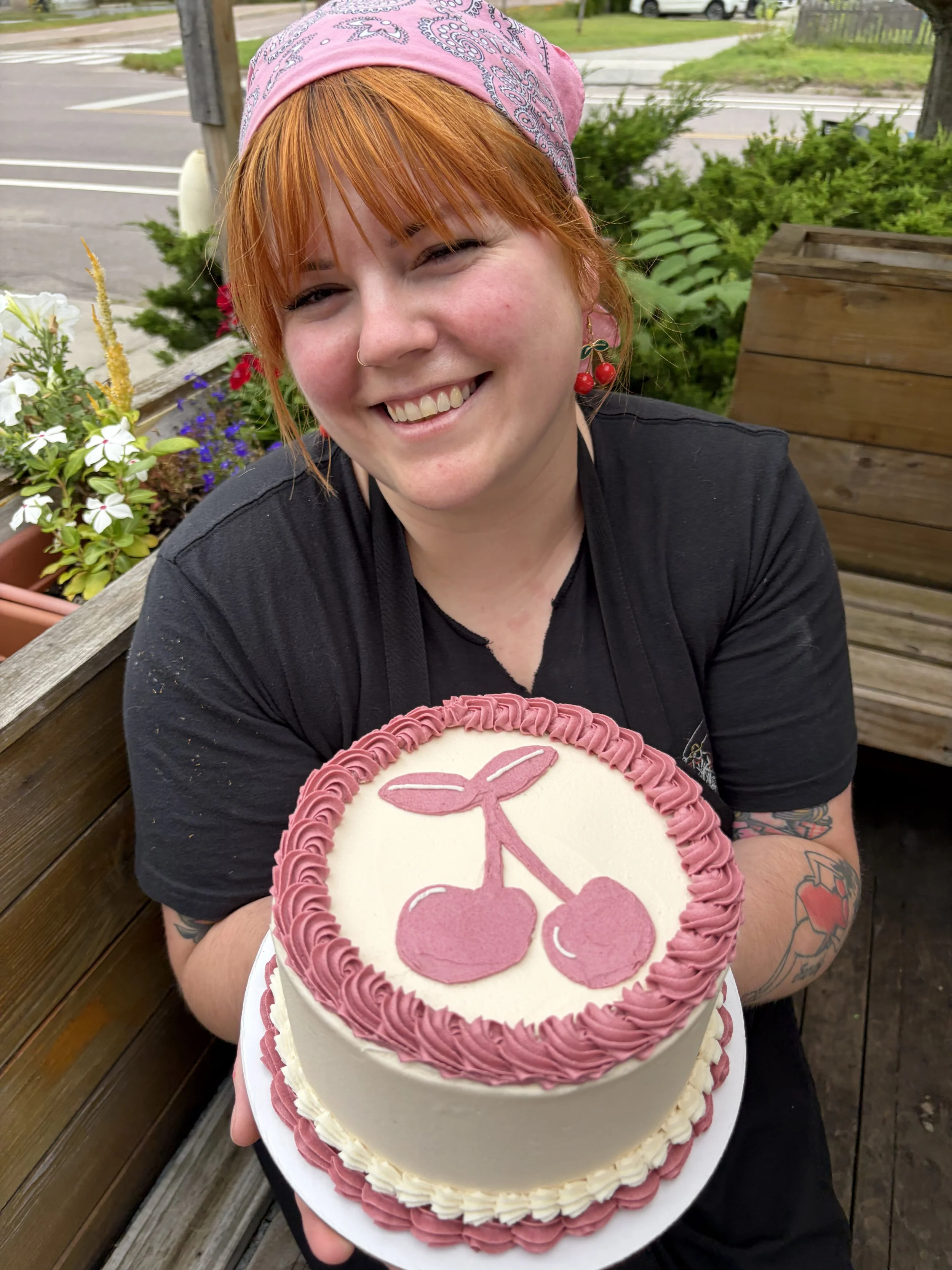 A woman with red hair wearing a pink bandana, earrings with cherries, and a black shirt, holding a birthday cake with a pink cherry design on top and pink and white frosting. She is smiling and sitting outdoors, with plants and a wooden bench behind her.