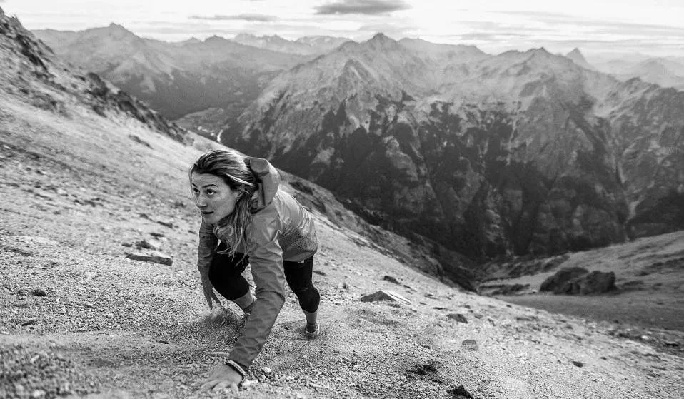 A woman in outdoor gear climbing up a steep mountain slope with rugged mountain peaks in the background.
