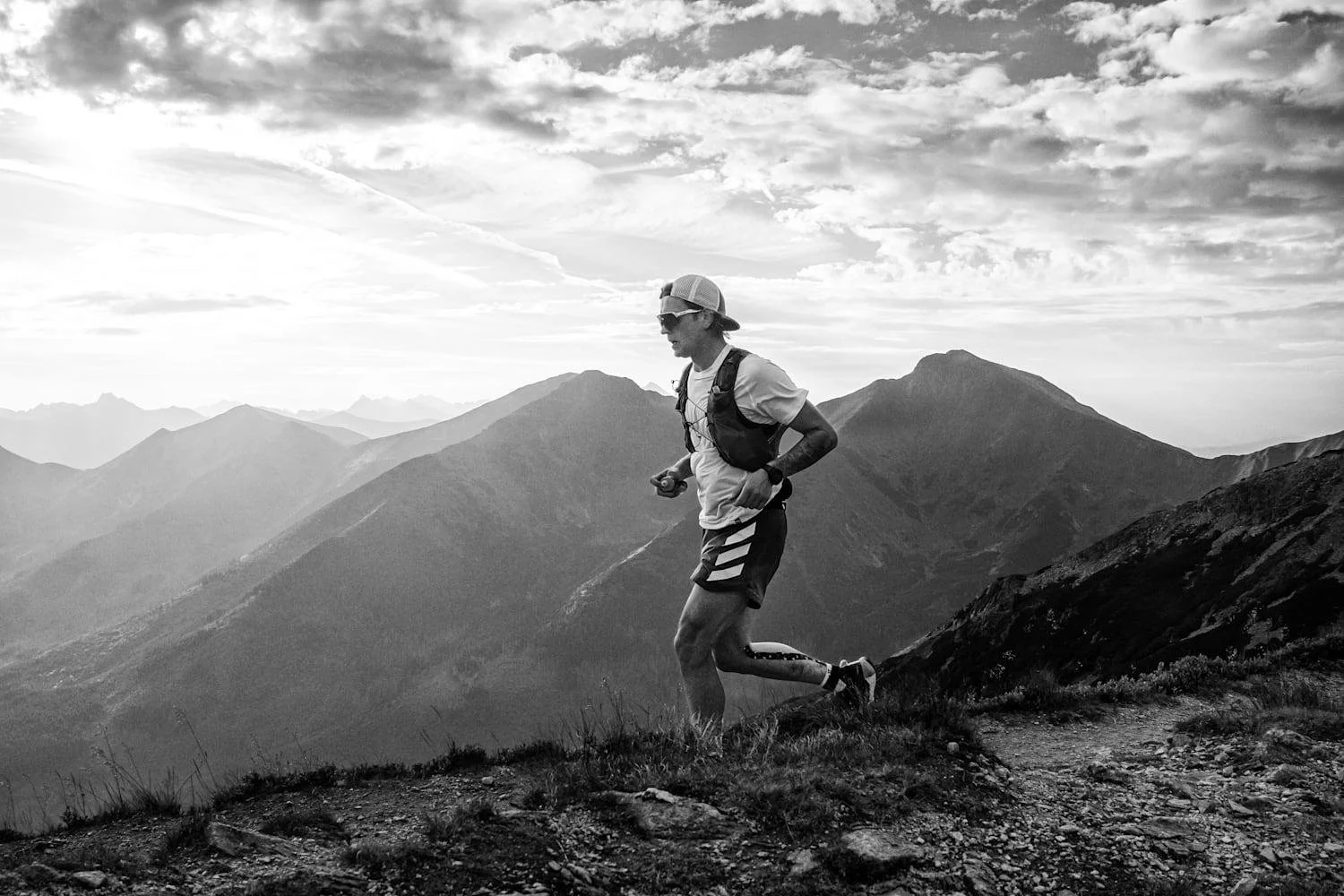 A person trail running on a mountain trail with mountain ranges and cloudy sky in the background in black and white.
