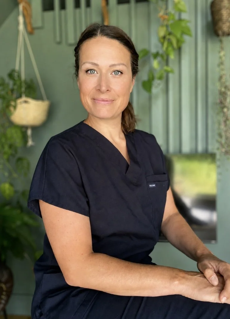 A woman with brown hair and blue eyes wearing a black uniform, seated indoors in a room with green walls, plants, and hanging planters, looking at the camera.