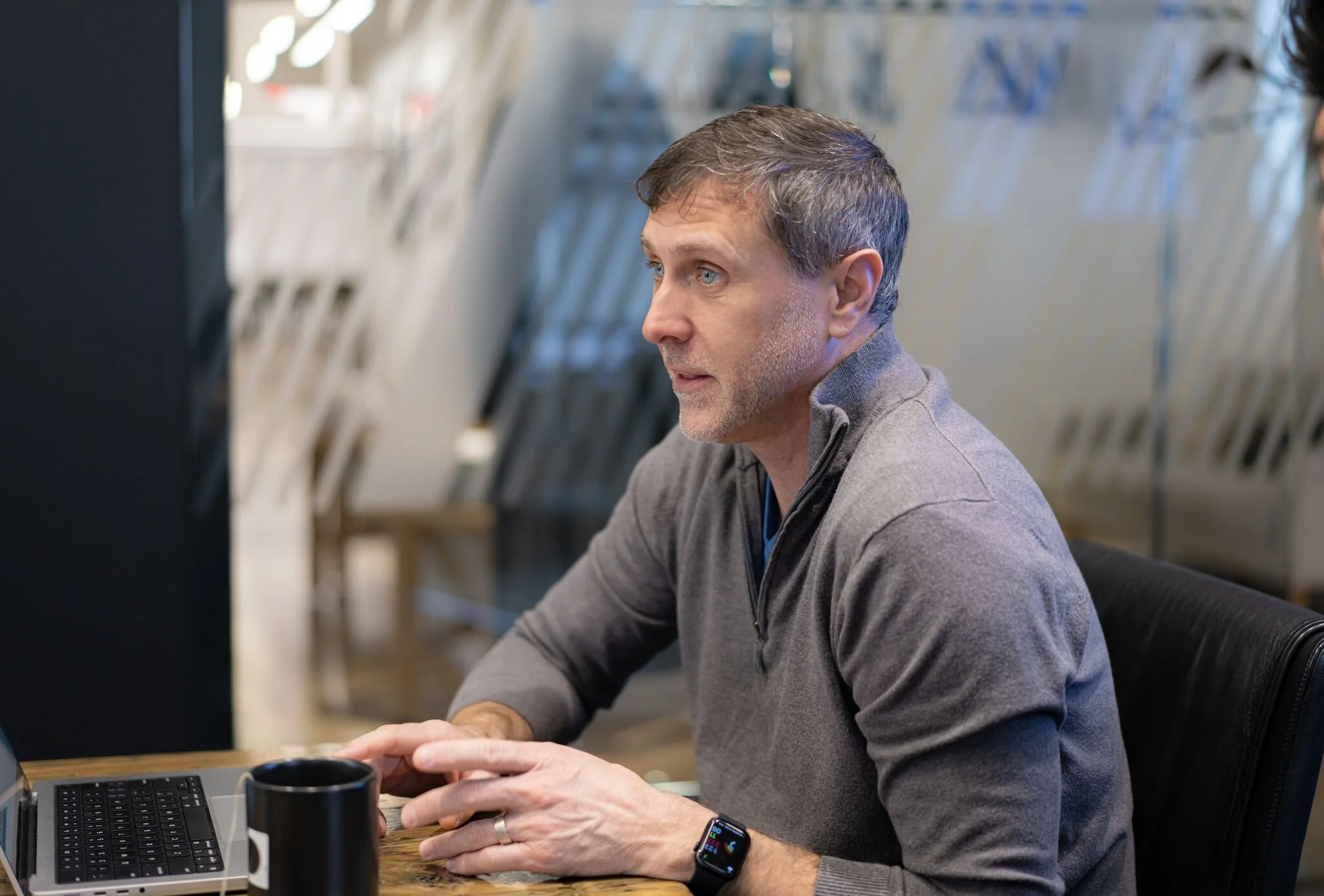 A man with gray hair and a beard wearing a gray zip-up jacket, sitting at a table with a laptop, coffee mug, and smartwatch, working in a modern office space with metallic wall decor.