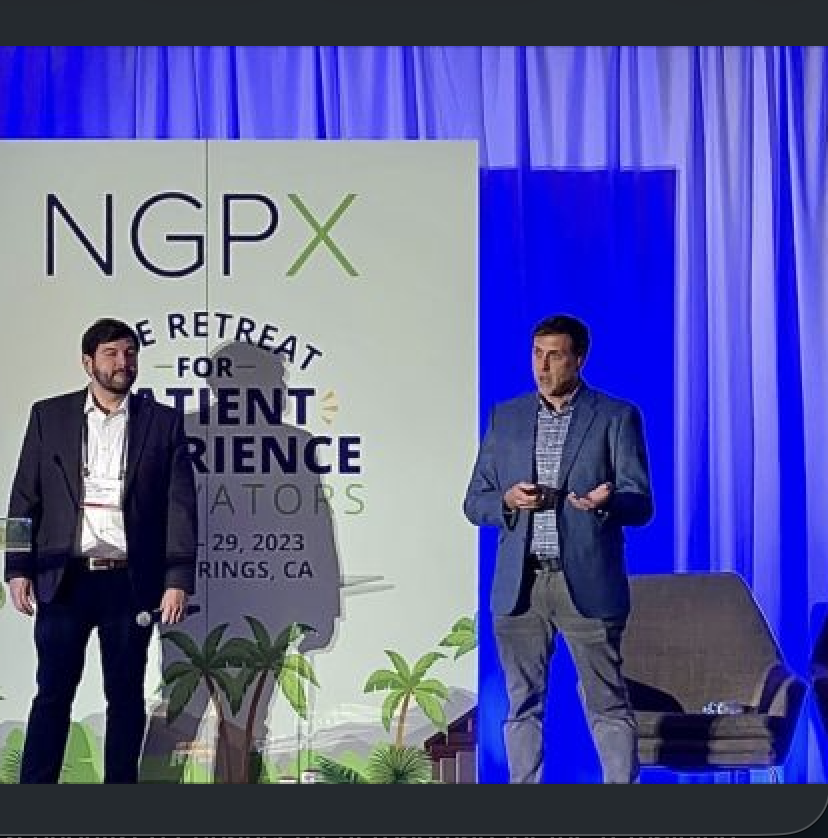 Two men in suits on a stage with a large screen behind them. The screen displays the text 'NGPX' and 'The Retreat for Content Creators' along with palm trees and a date, November 29, 2023, in RINGS, CA. The stage has blue curtains.