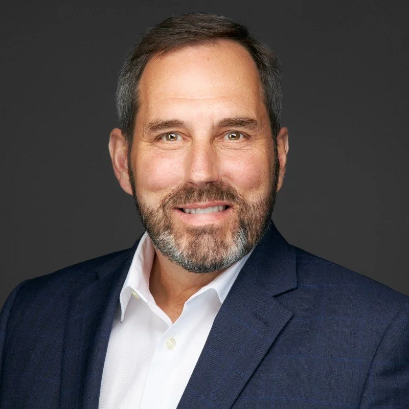 Professional headshot of a middle-aged man with a beard, wearing a dark suit and white shirt, smiling against a dark background.