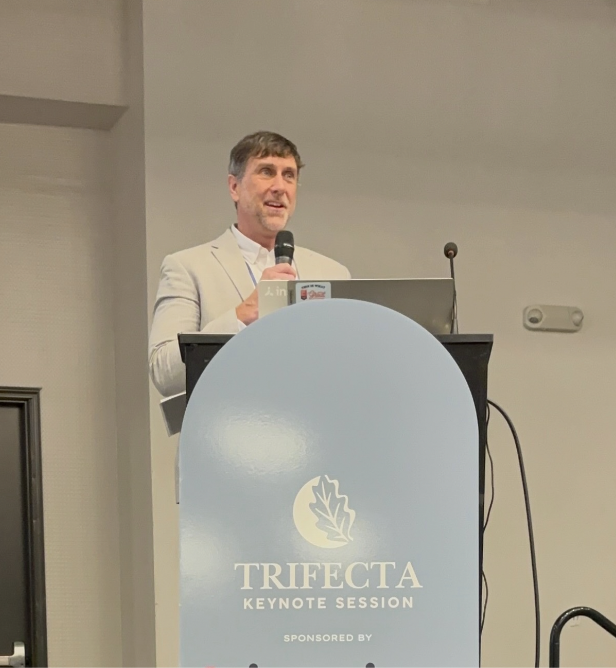A man in a beige suit standing behind a podium with a laptop, speaking into a microphone at the Trifecta Keynote Session, sponsored by .